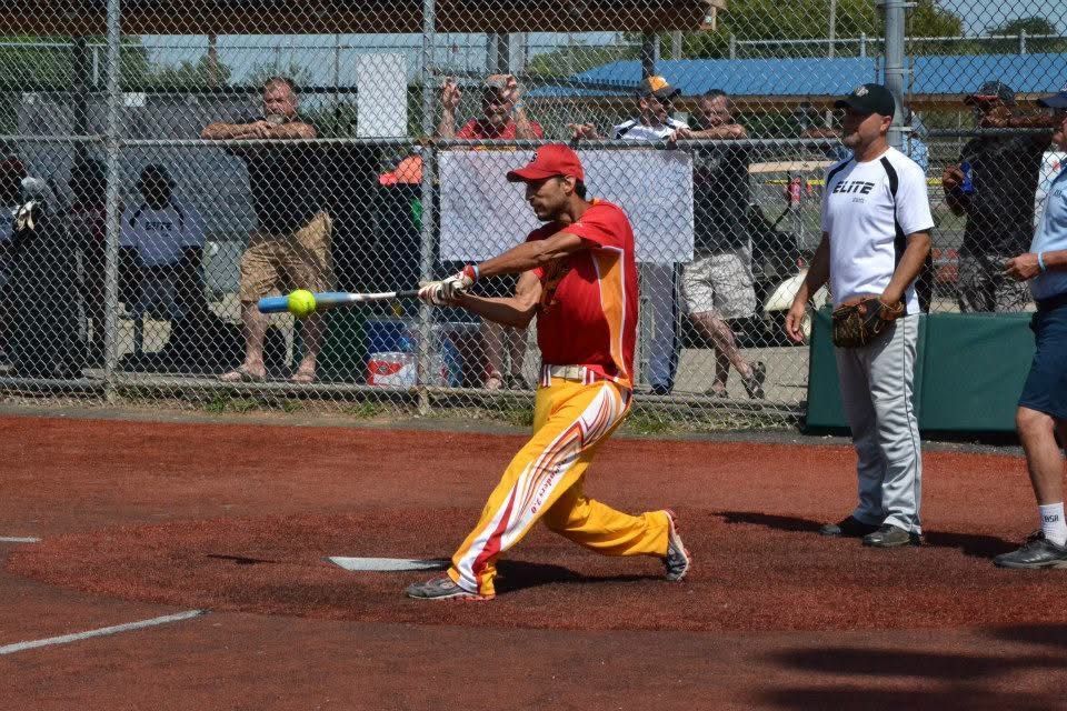 Softball batter in red swings at a pitch on a dirt field, with umpire and spectators behind the fence.