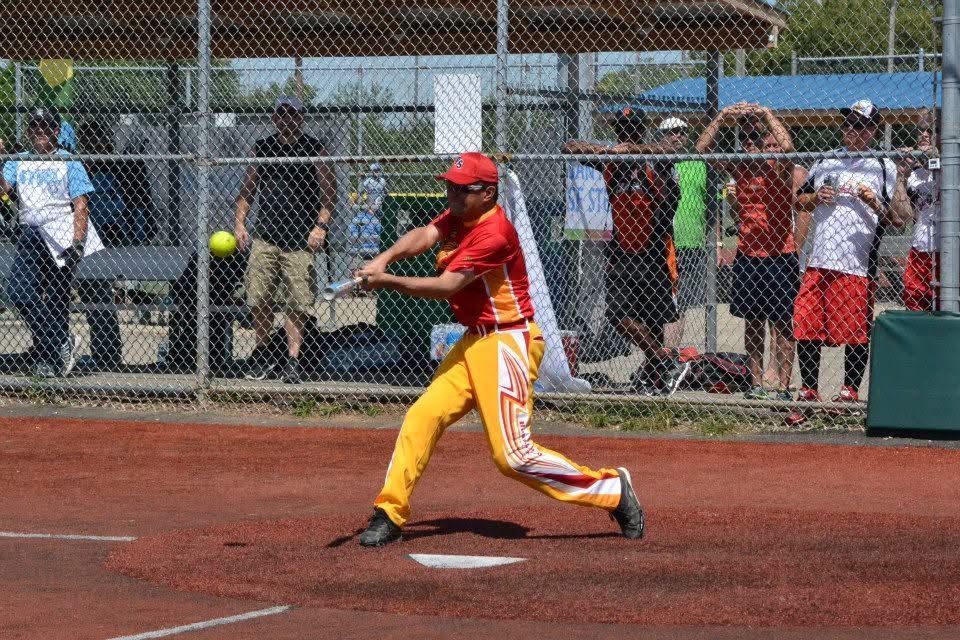 Softball batter in red and yellow uniform swings at a pitch on a dirt field, spectators behind a fence.