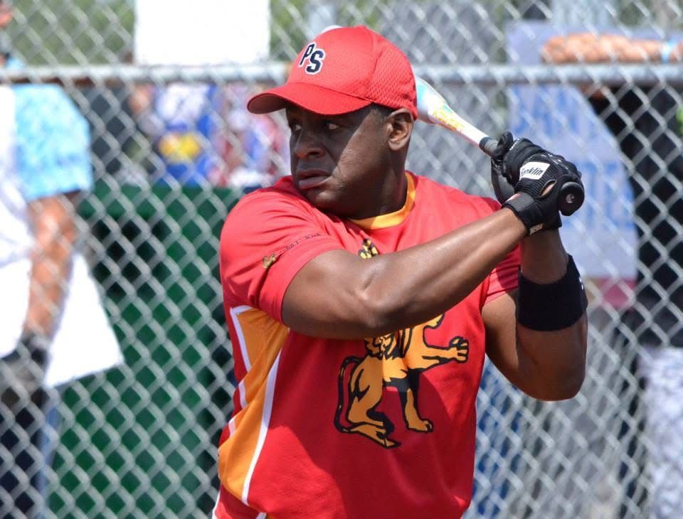 Baseball player in red uniform and cap, holding a bat in a batting stance near a chain-link fence.