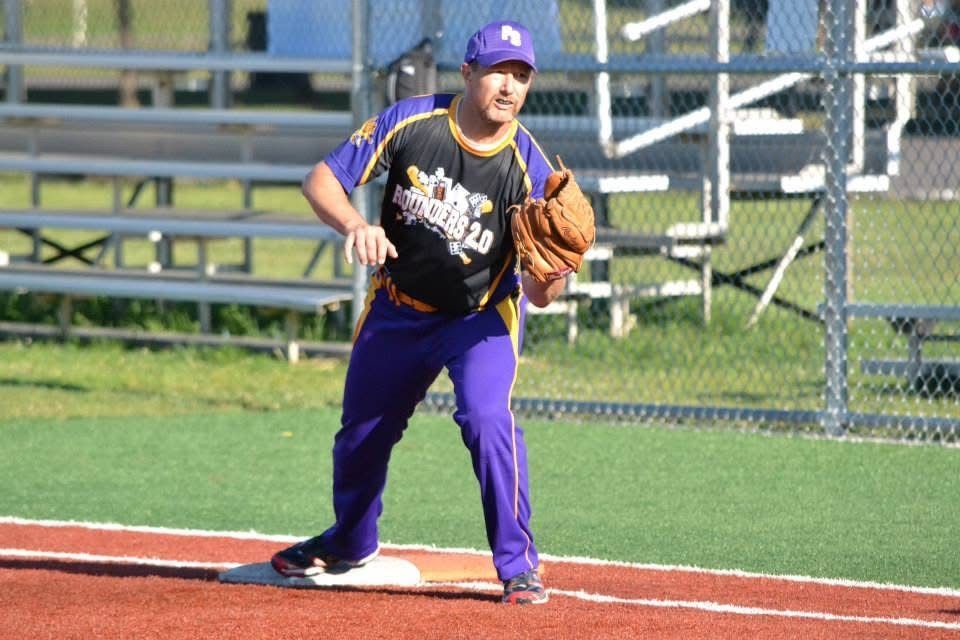 Baseball player in purple uniform and cap fields a ball near first base on an outdoor field
