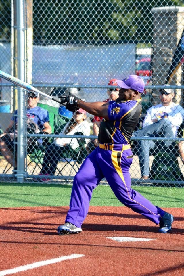 Softball batter in purple uniform swinging at a pitch on a field, with chain-link fence and spectators behind.