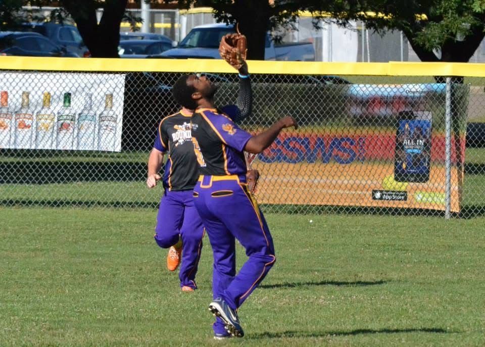 Two softball players in purple uniforms leap for a catch near the outfield fence.