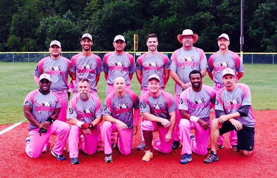Group of softball players in pink uniforms posing on a field, with a tree line in the background.