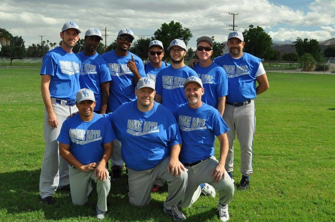 Nine people in blue shirts pose on a grassy field with trees and buildings in the background.