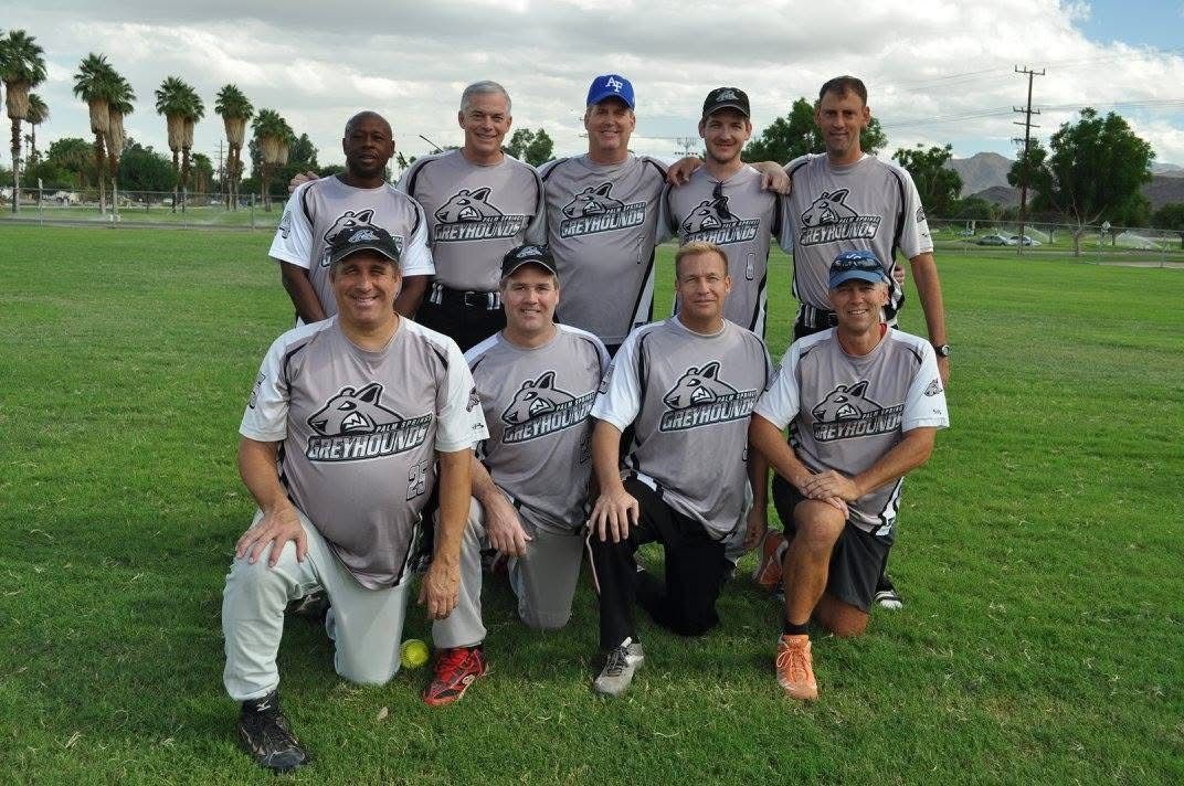 Group of eight people in gray jerseys posing on a grassy field, with trees and a cloudy sky behind them.