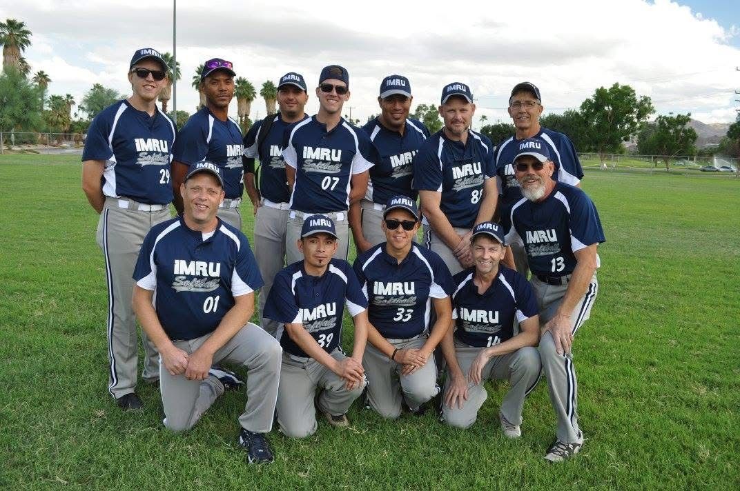 Baseball team posing on a grassy field, wearing navy jerseys and gray pants, smiling at the camera.