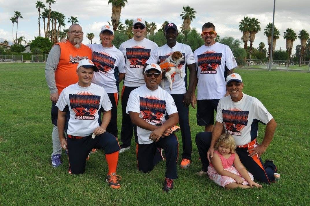 Group of athletes posing on a grassy field, wearing white and orange sports shirts and caps, with palm trees behind them