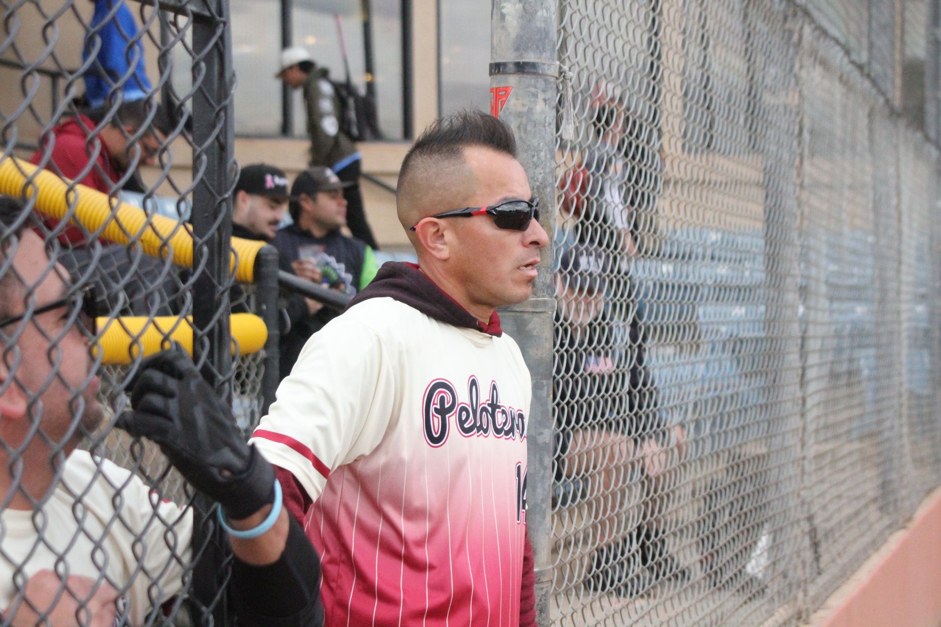 Baseball player in white and pink uniform standing by the dugout fence, wearing sunglasses and batting gloves