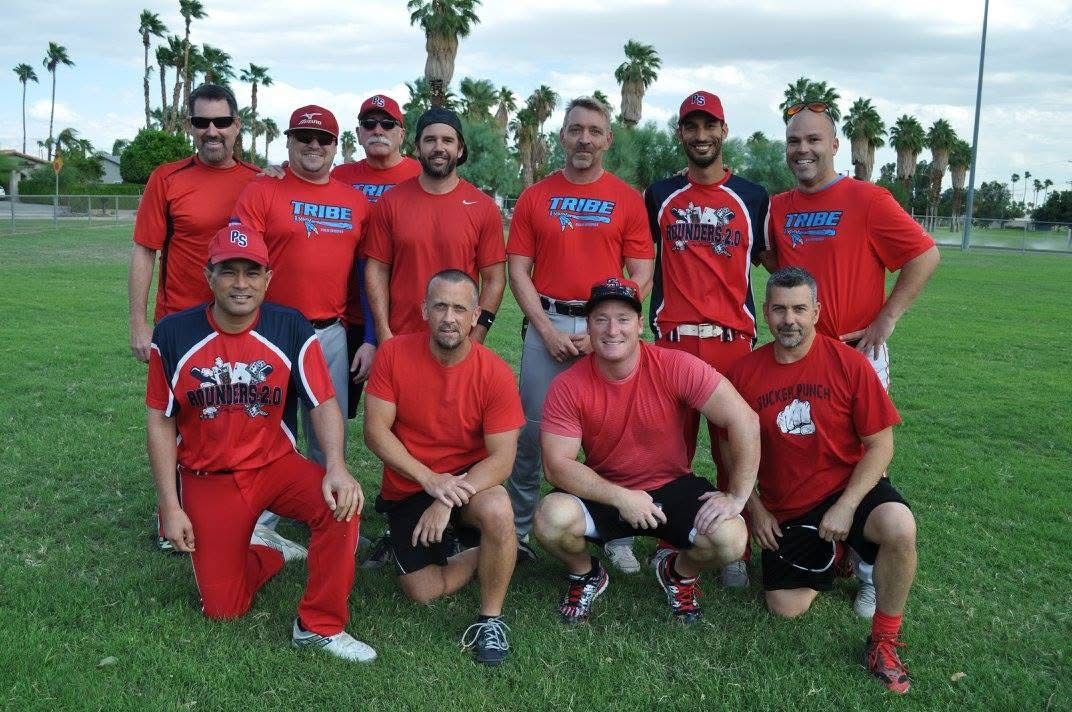 Group of men in red sports shirts posing on a grassy field with palm trees in the background