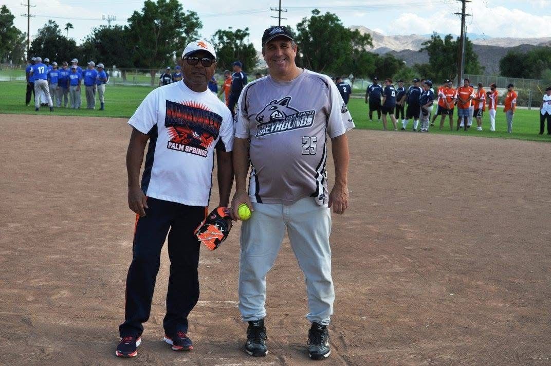 Two people posing on a softball field, with teams lined up in the background.