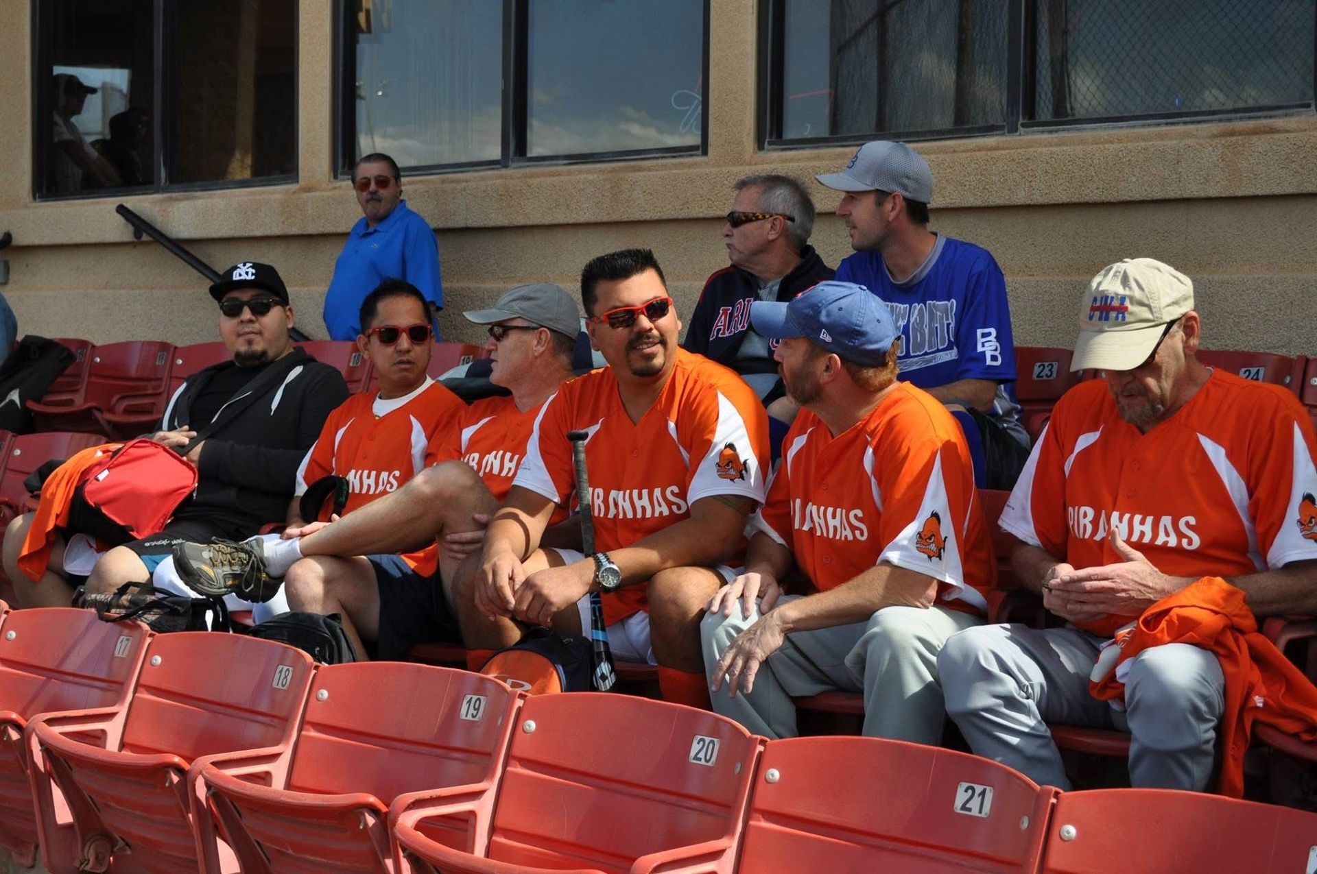 Baseball fans in orange and red jerseys sit in stadium seats, chatting and watching the game.
