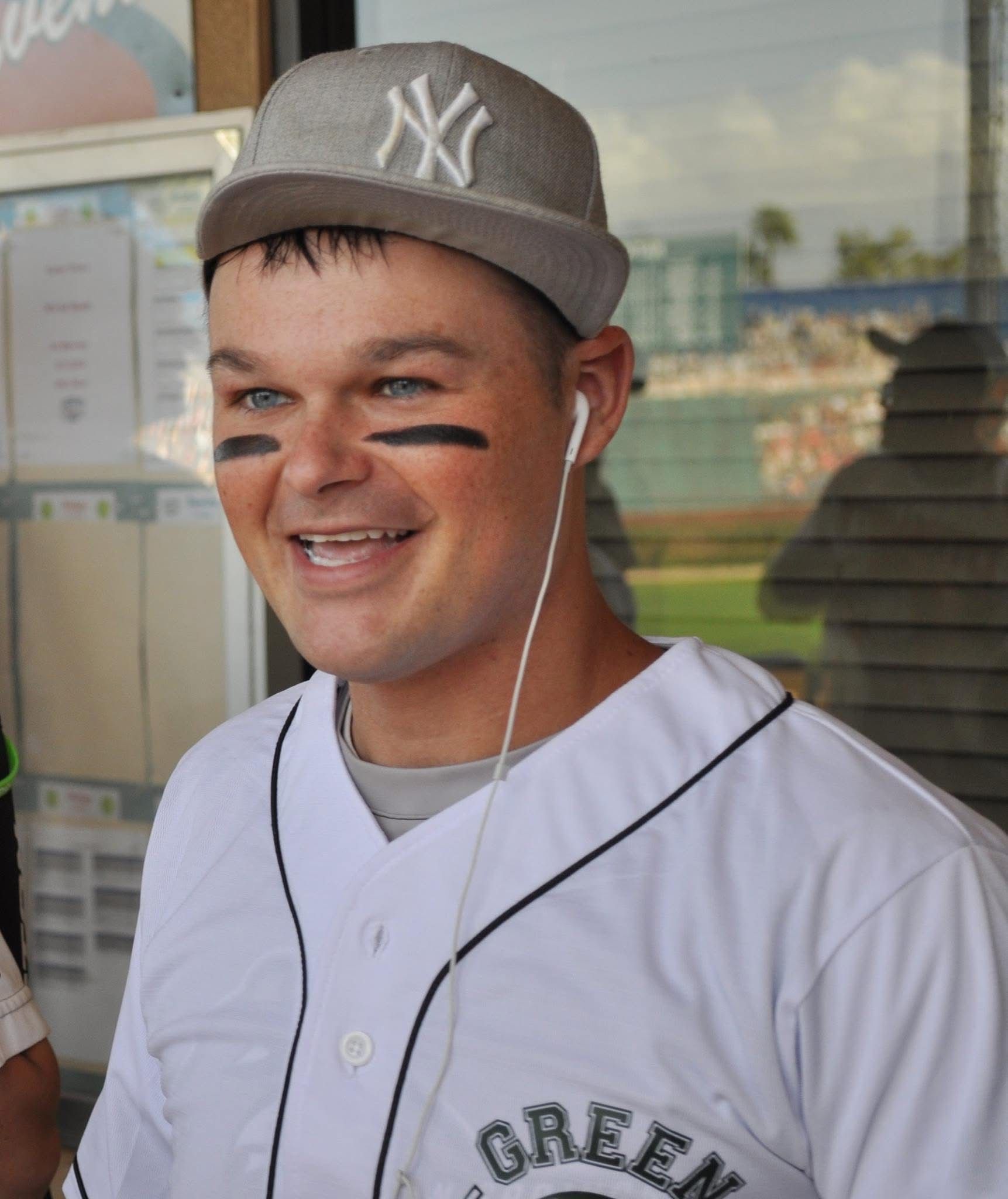 Smiling baseball player in white jersey and gray cap, with black eye paint and earbuds, standing outdoors