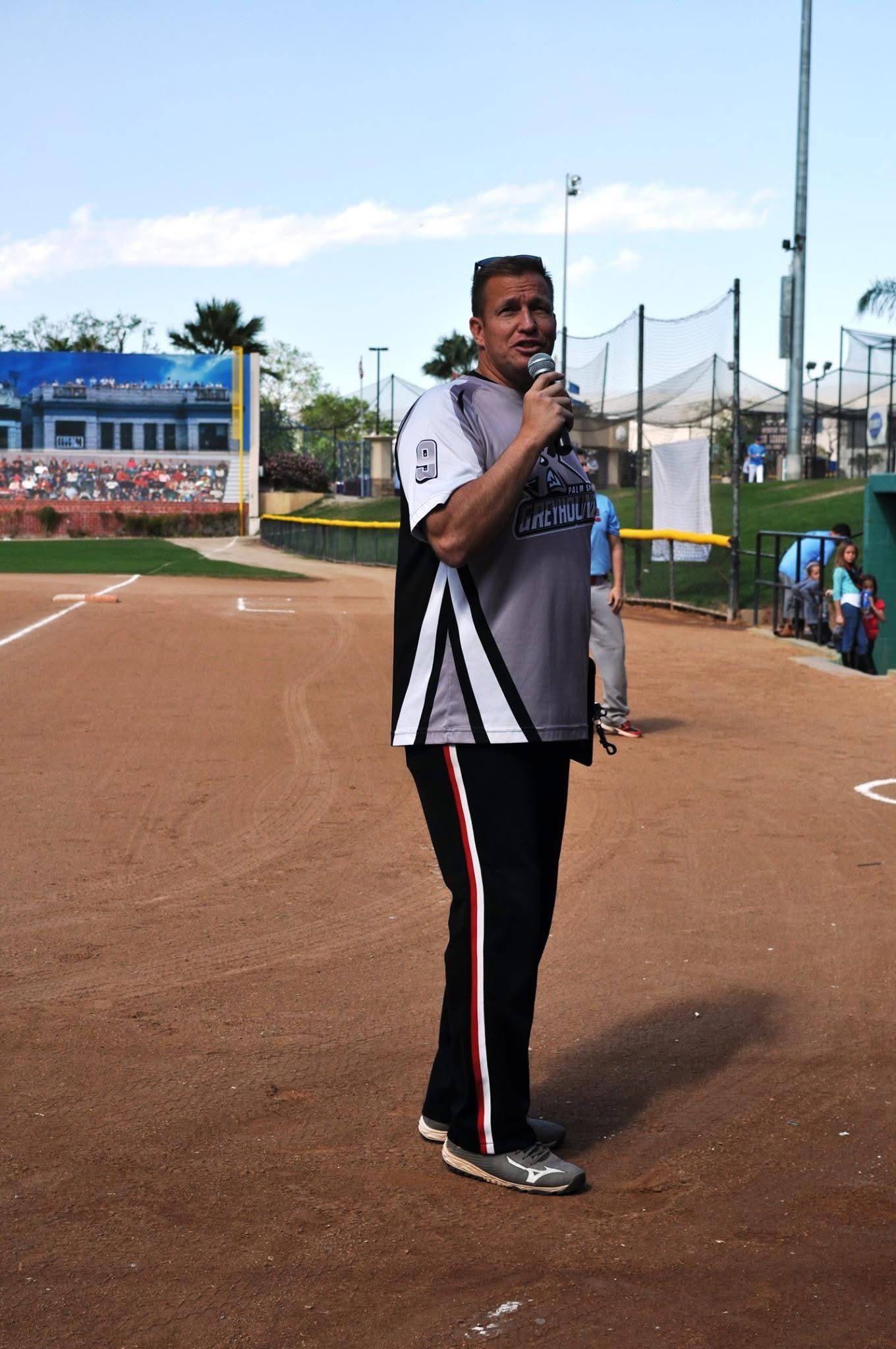 Man standing on a track holding a microphone at a sports field, with bleachers and equipment in the background