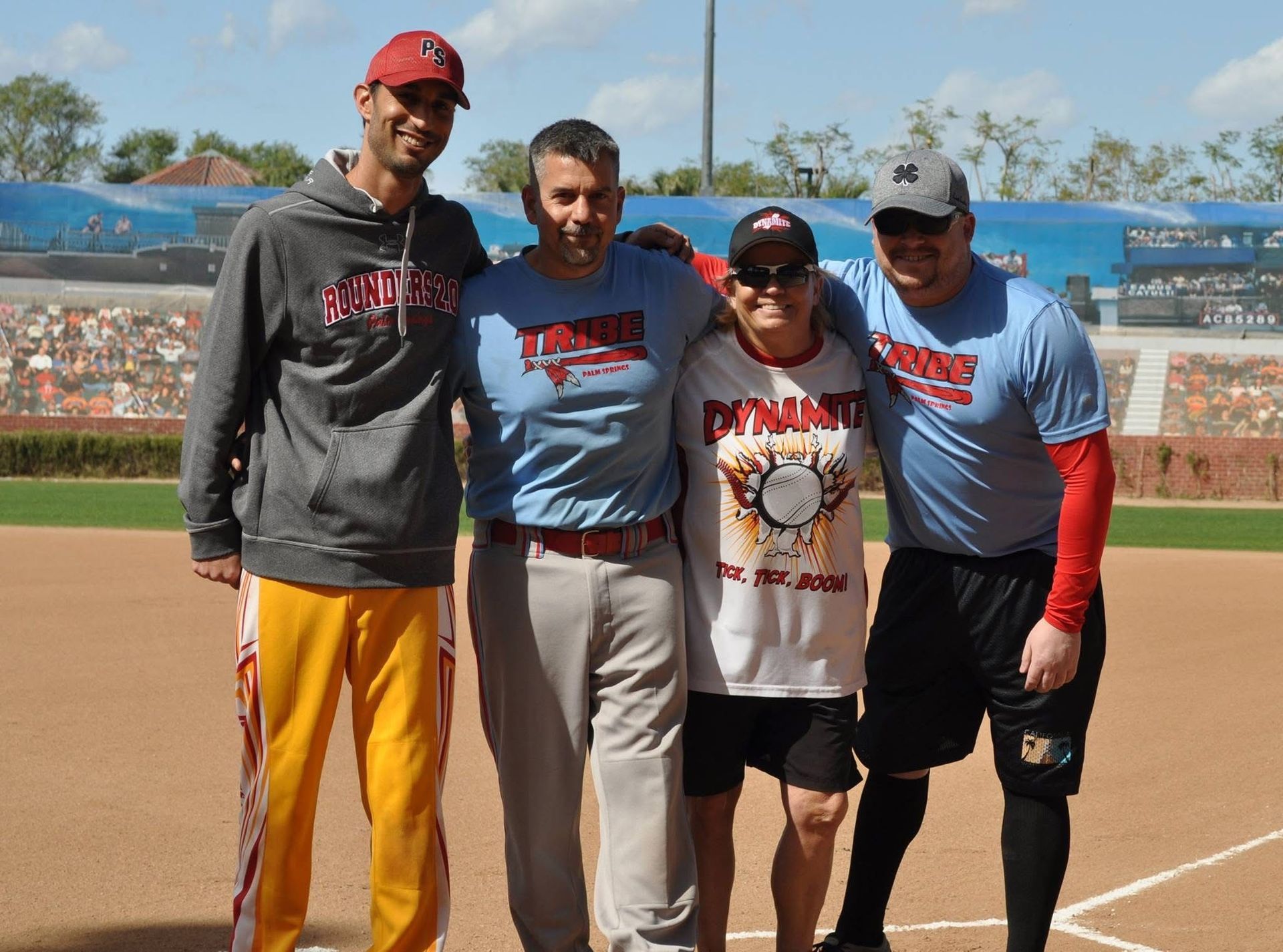 Four people posing together on a baseball field, wearing team shirts and caps, with stadium stands in the background