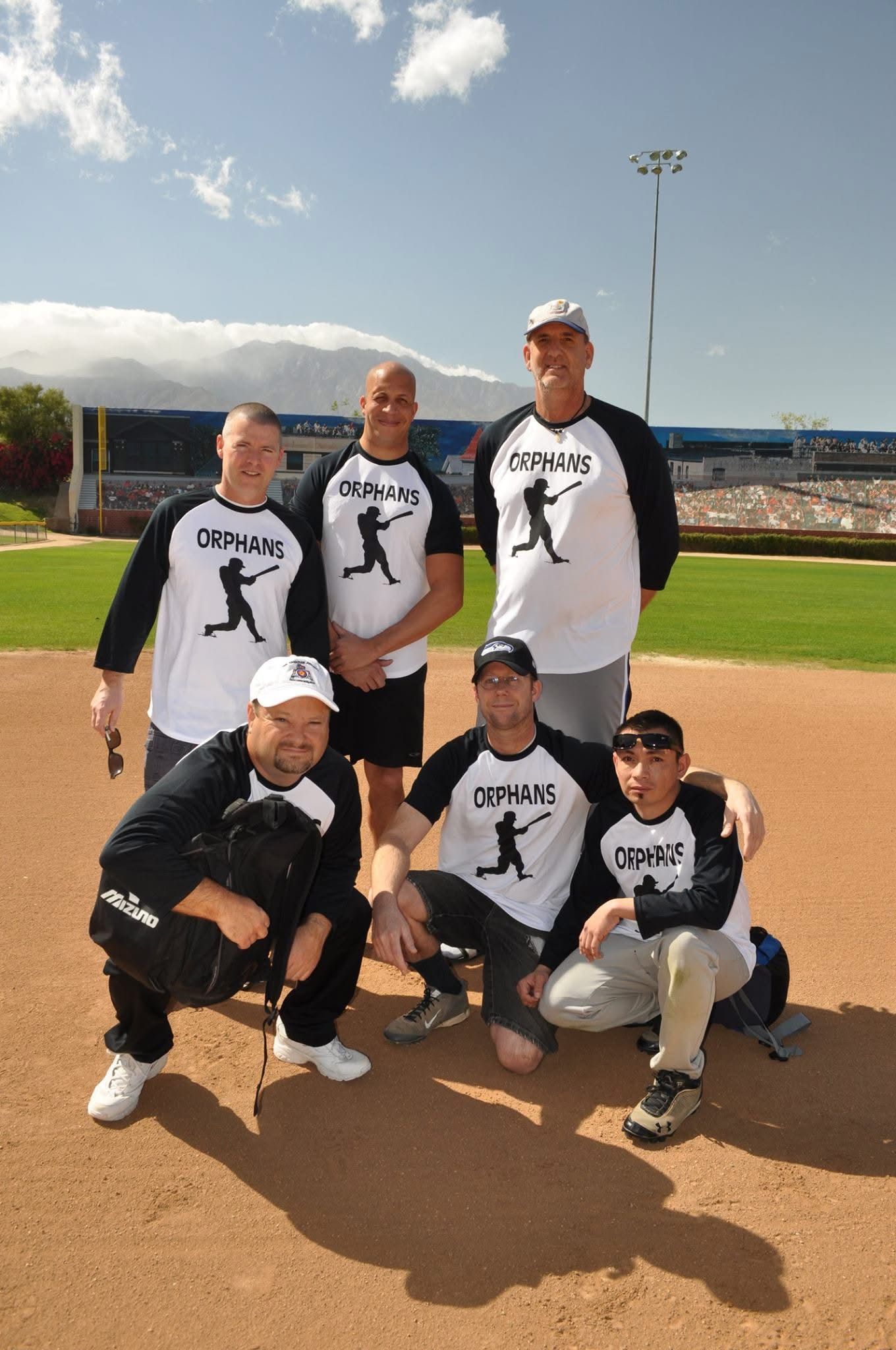 Six people in matching baseball shirts pose on a field under a blue sky.