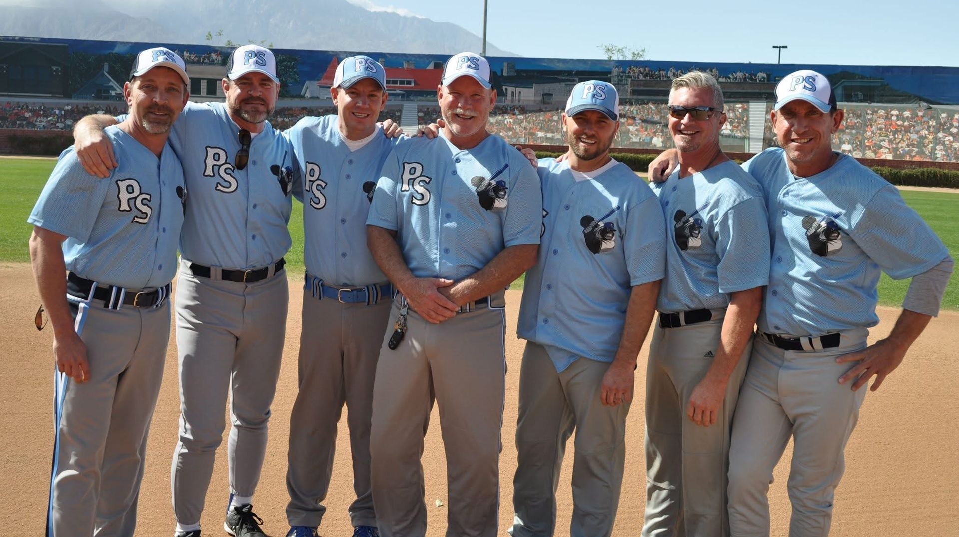 Seven baseball players in light blue uniforms pose together on a field, smiling at the camera.