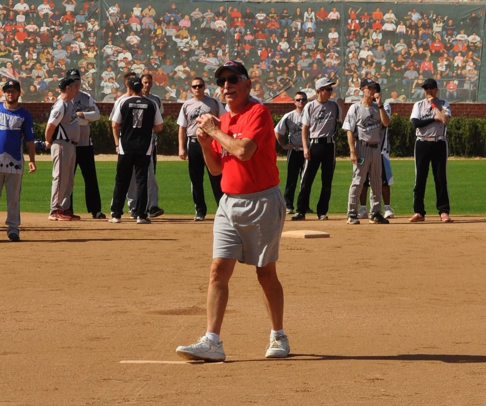 Man in red shirt and gray shorts standing on a baseball field with players lined up behind him