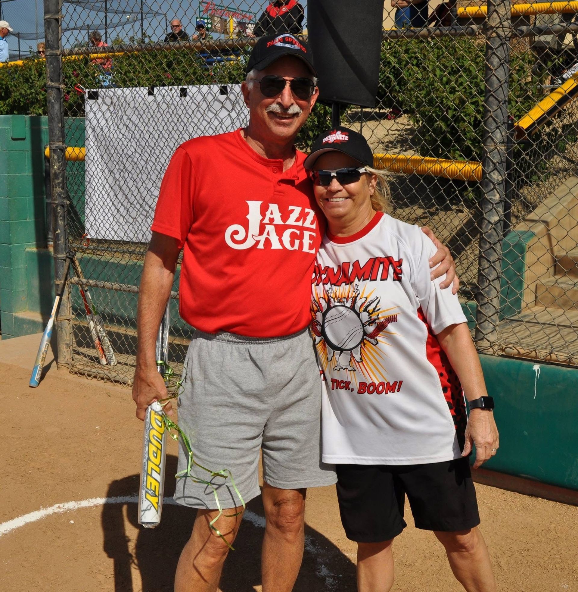 Two people posing on a tennis court, smiling and holding rackets in front of a chain-link fence.