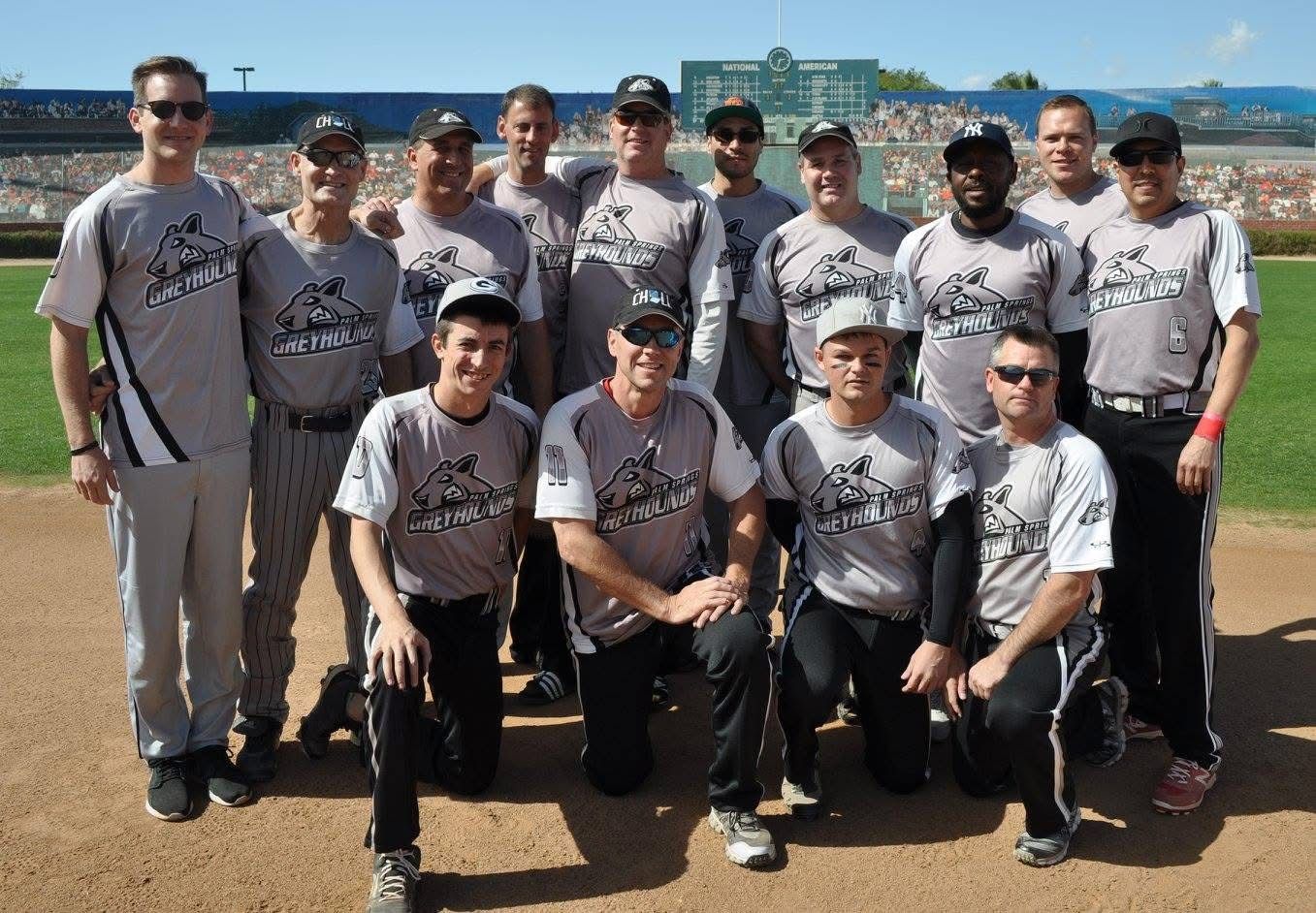 Baseball team posing on a field in gray uniforms, with a stadium and blue sky in the background.