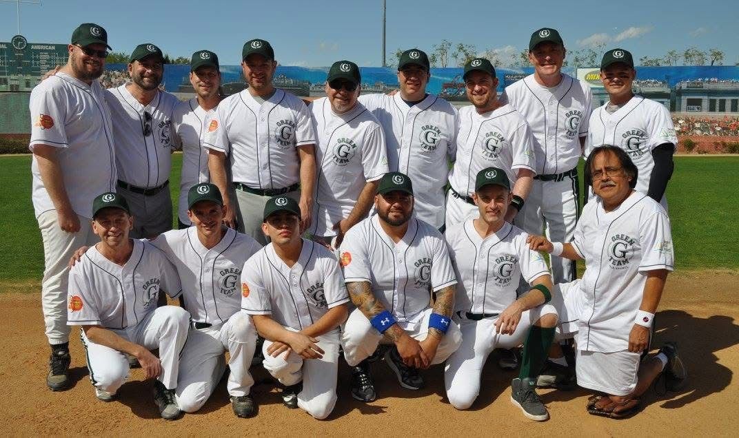 Baseball team posing together in white uniforms on a field, some kneeling in front, with a water and grass background