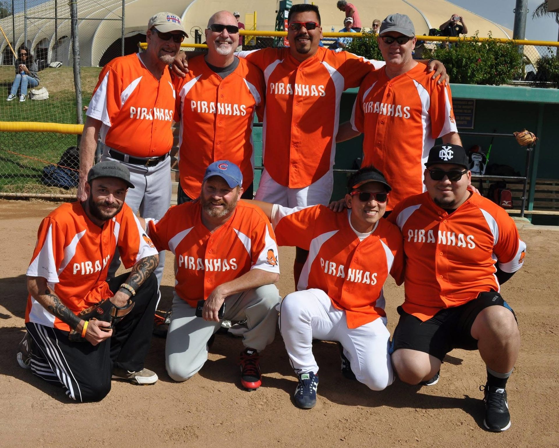 Eight softball players in orange jerseys pose together on a dirt field, smiling for a team photo.