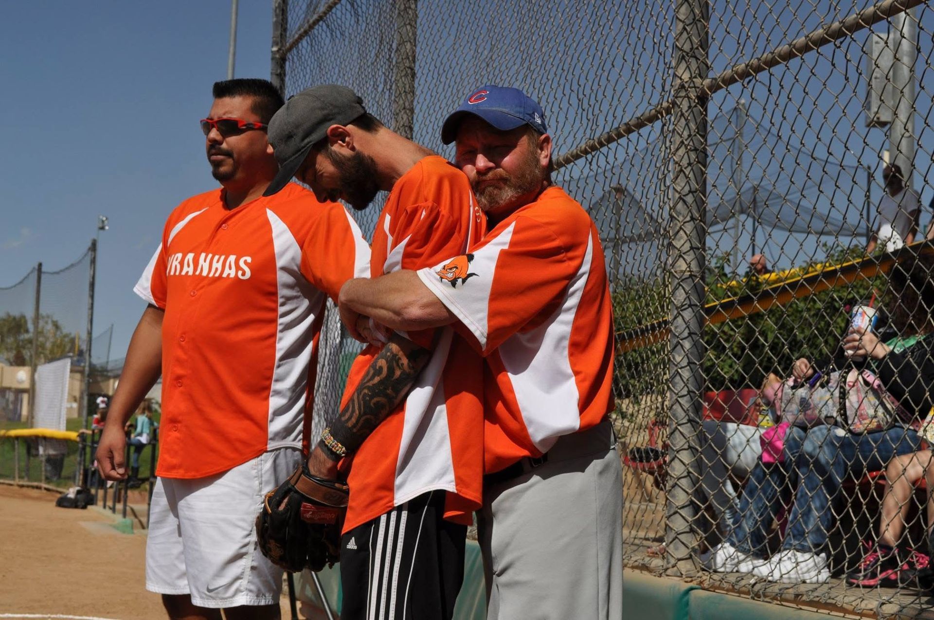Three men in orange sports shirts stand by a chain-link fence at a baseball field.