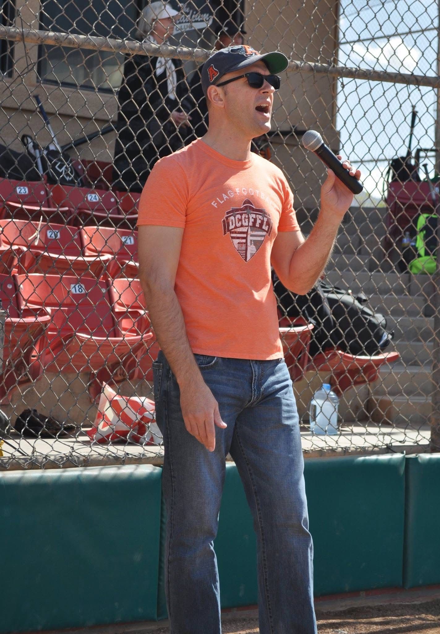 Man in orange shirt speaking into a microphone beside a baseball field dugout
