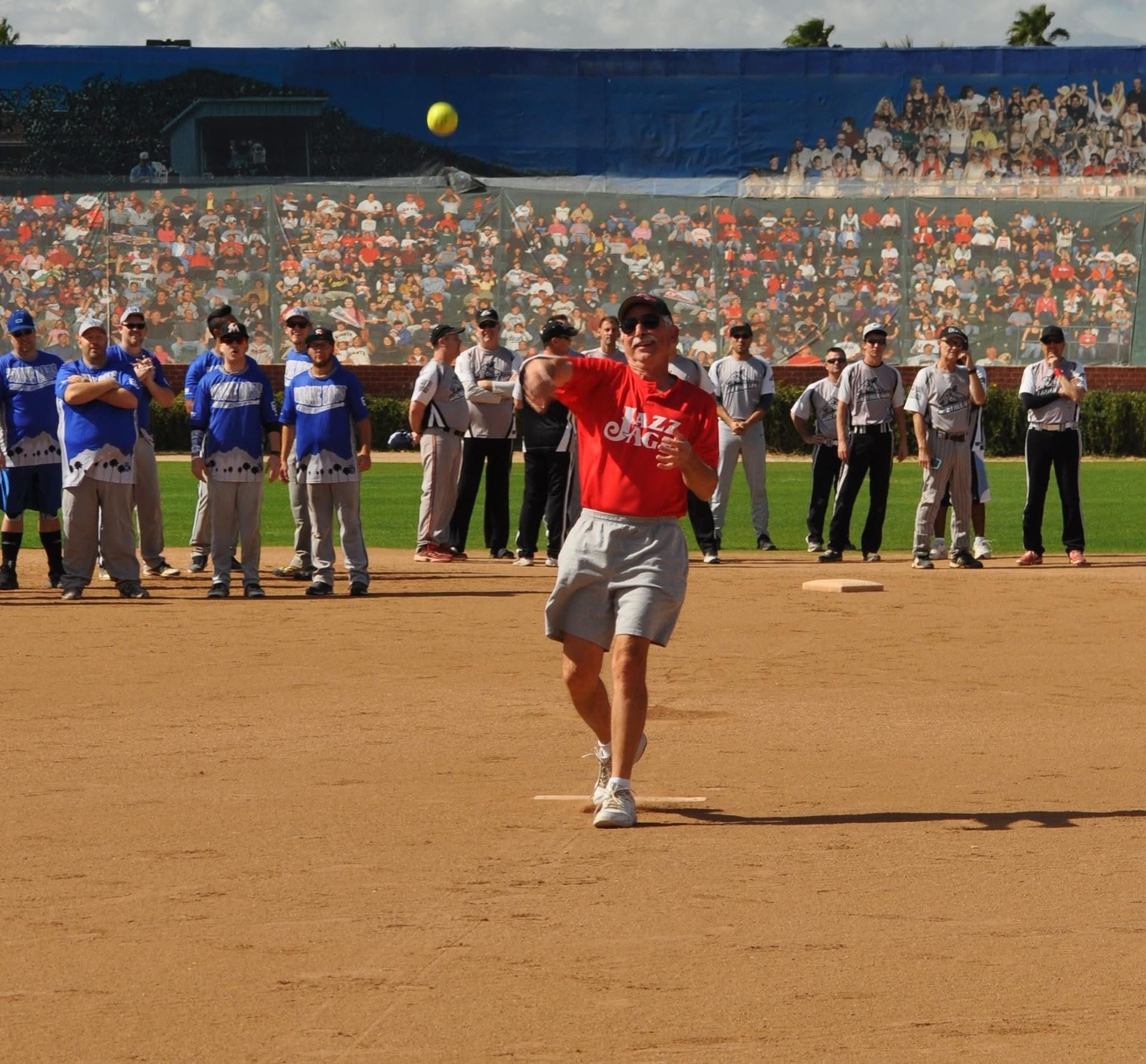 Softball pitcher in red shirt throws a pitch on a dirt field during a game, with players and crowd in the background