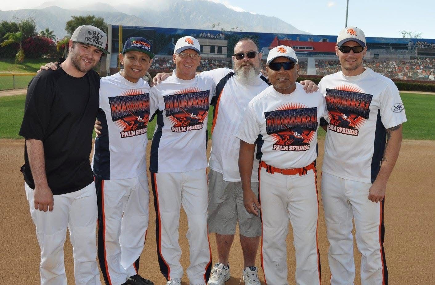 Six baseball players and a coach pose on a field, wearing white uniforms and black shirts under bright daylight.