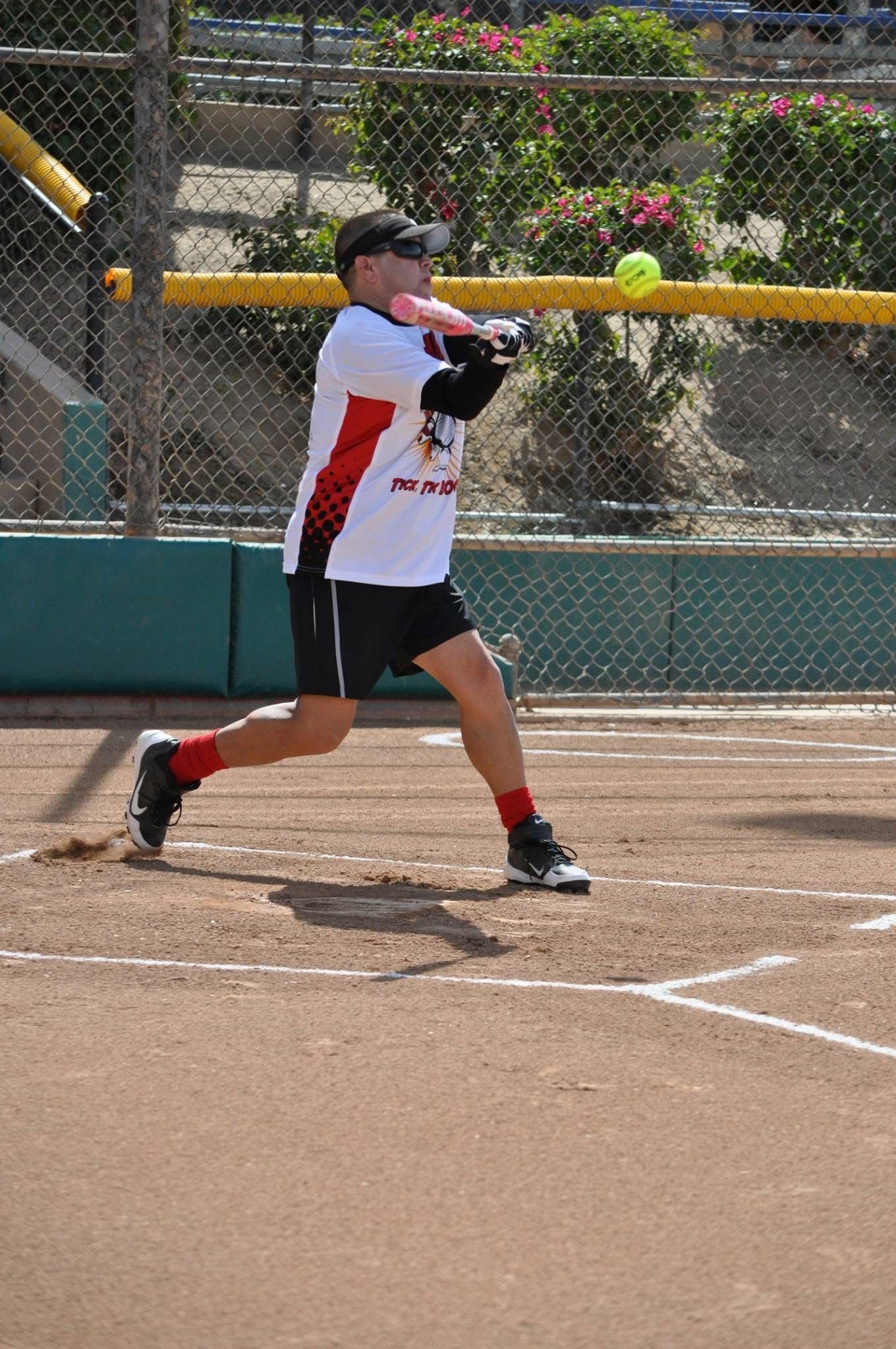 Softball player swinging at a ball on a dirt field near the fence