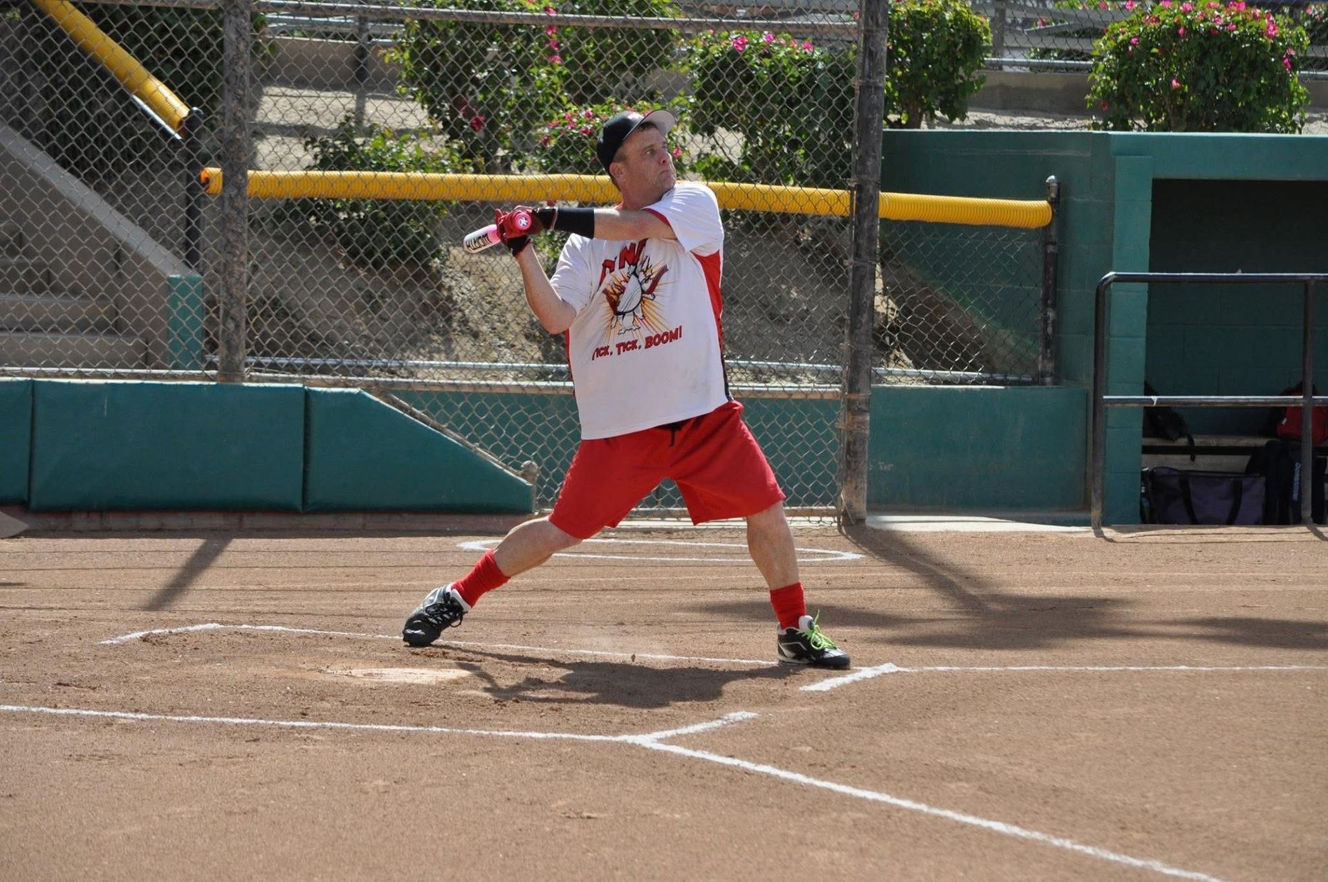 Softball batter in white and red uniform swinging a pink bat at home plate on a dirt field