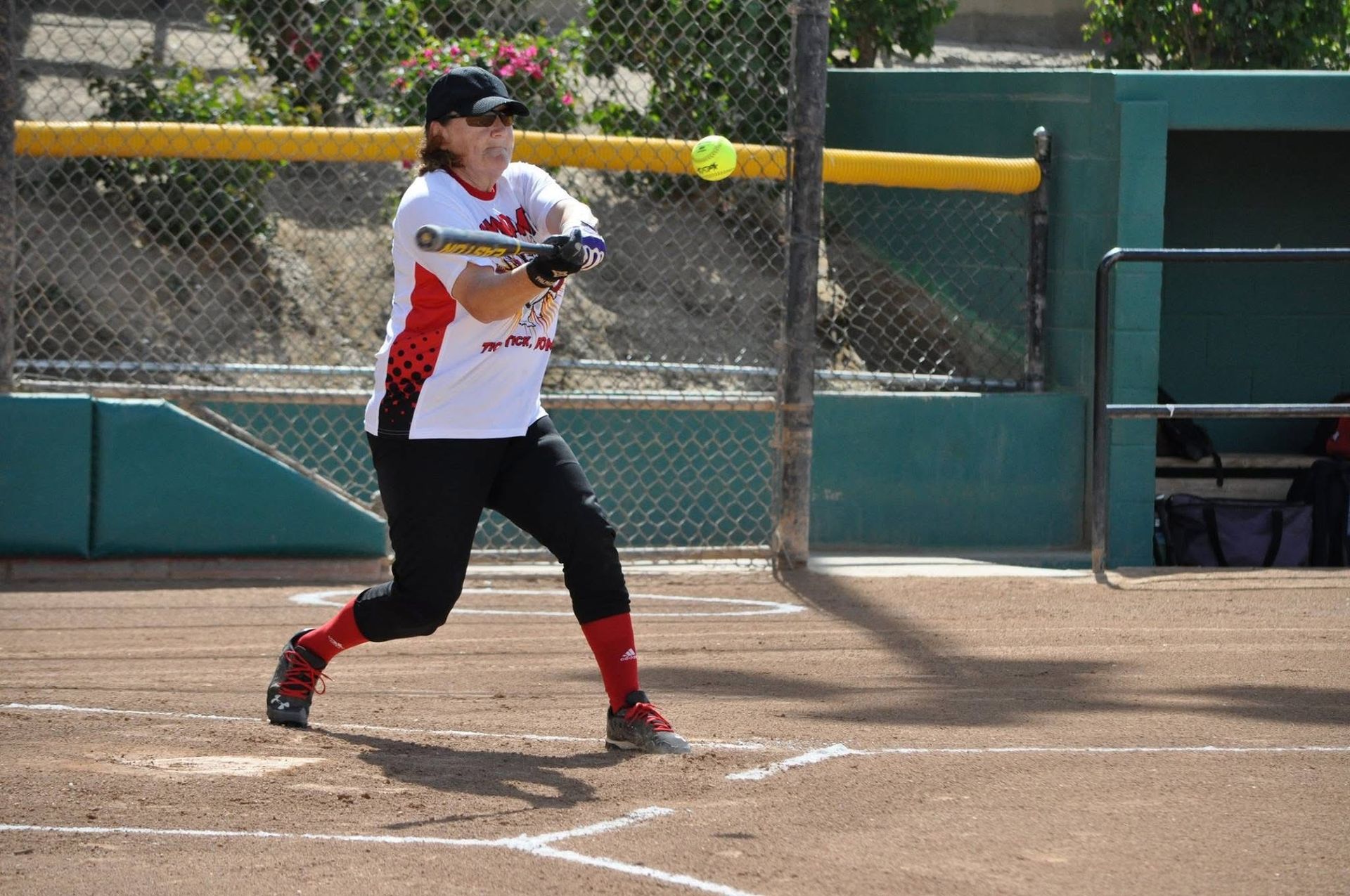 Softball batter in white and red uniform swinging at home plate on a dirt field