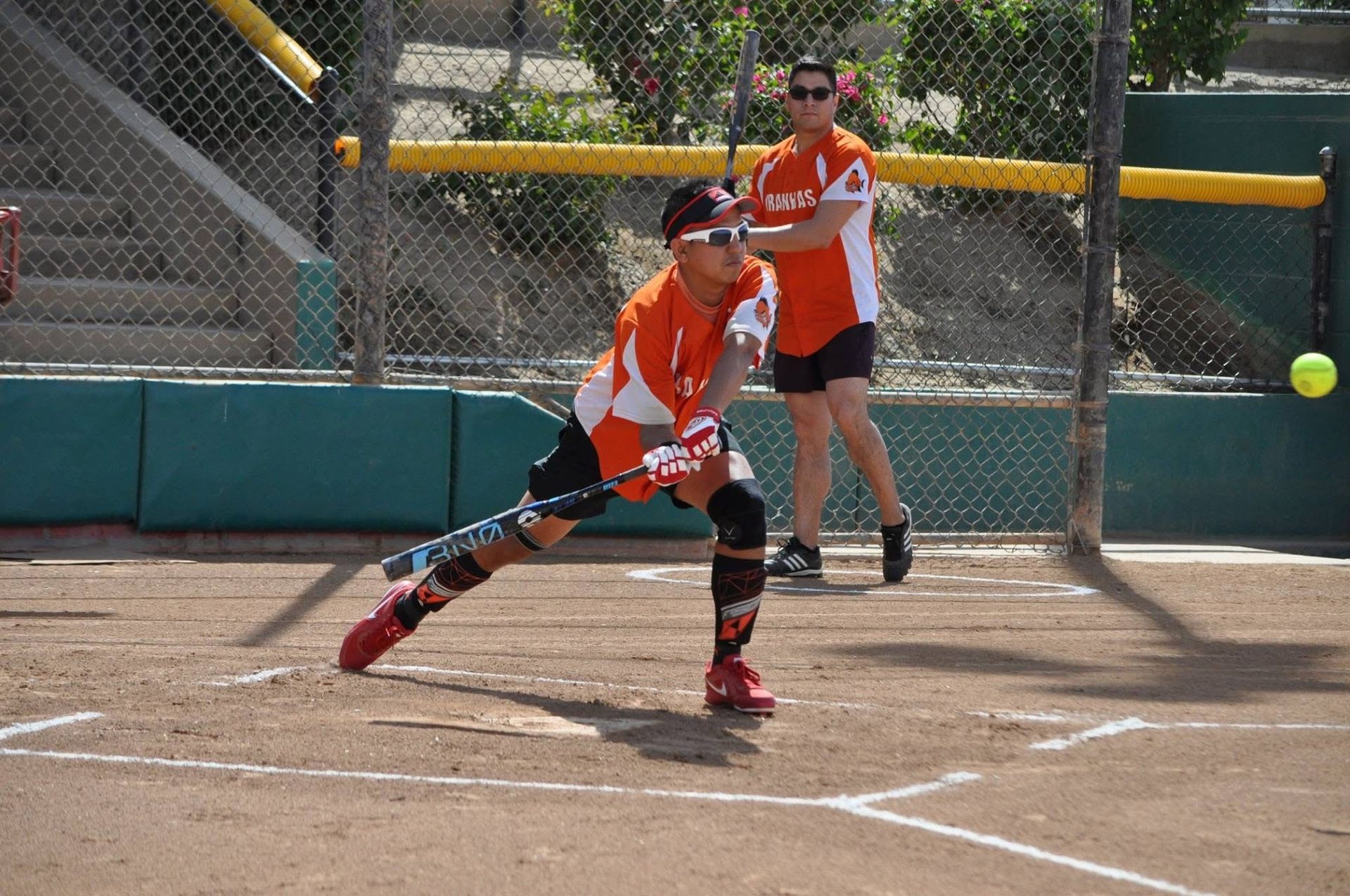 Softball player in orange uniform swings at pitch on a dirt field, with catcher behind the plate.