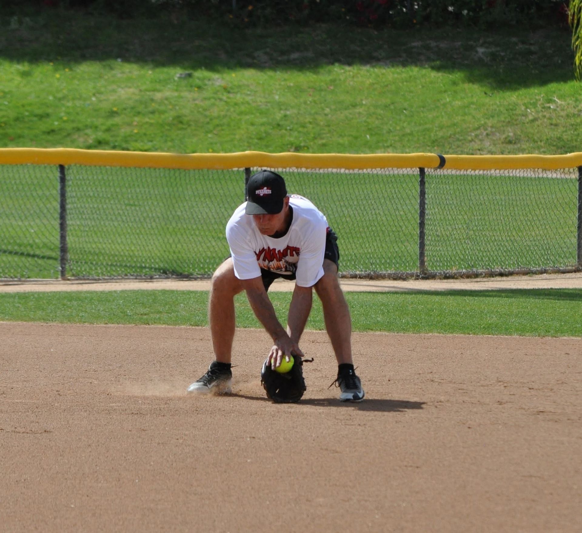 Softball player crouches over a ball on the infield, with a green outfield and fence behind.