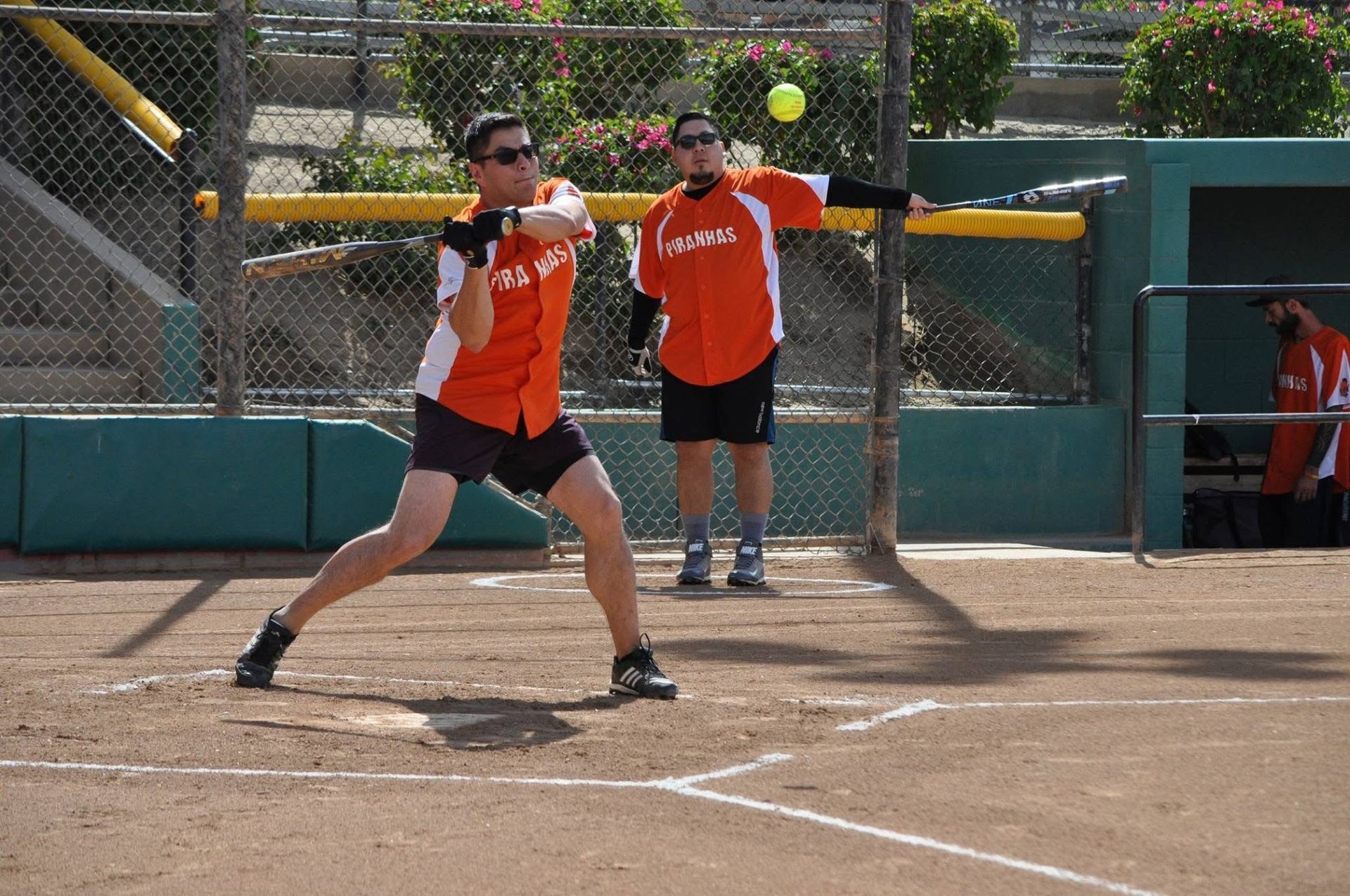 Softball batter in orange swings at a pitch on a dirt field, with catcher and umpire behind her.