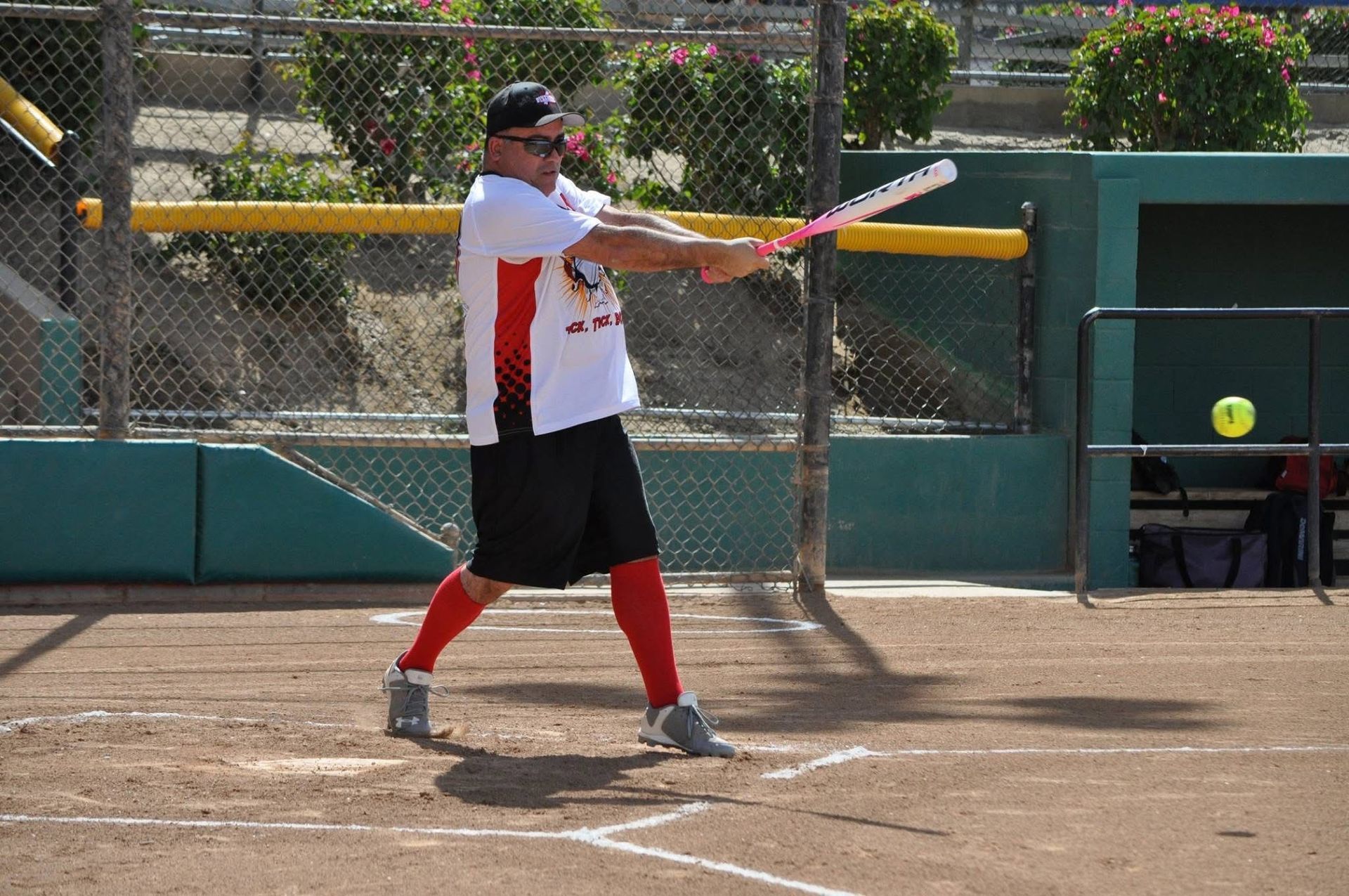 Softball player in white shirt and red socks swings a bat on a dirt field, with a ball in midair nearby
