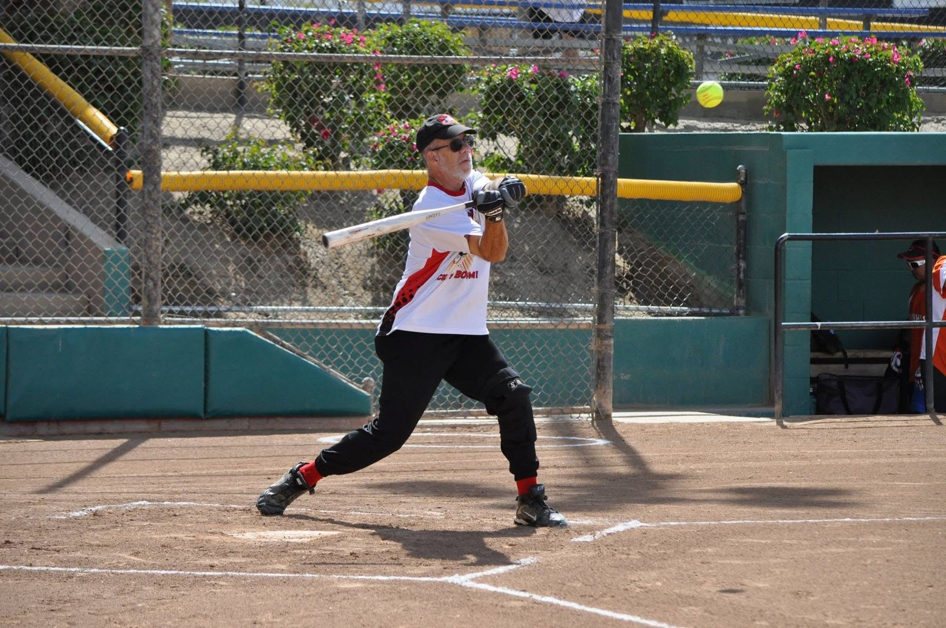 Softball player in white and black swings a bat at home plate on a sunny field.