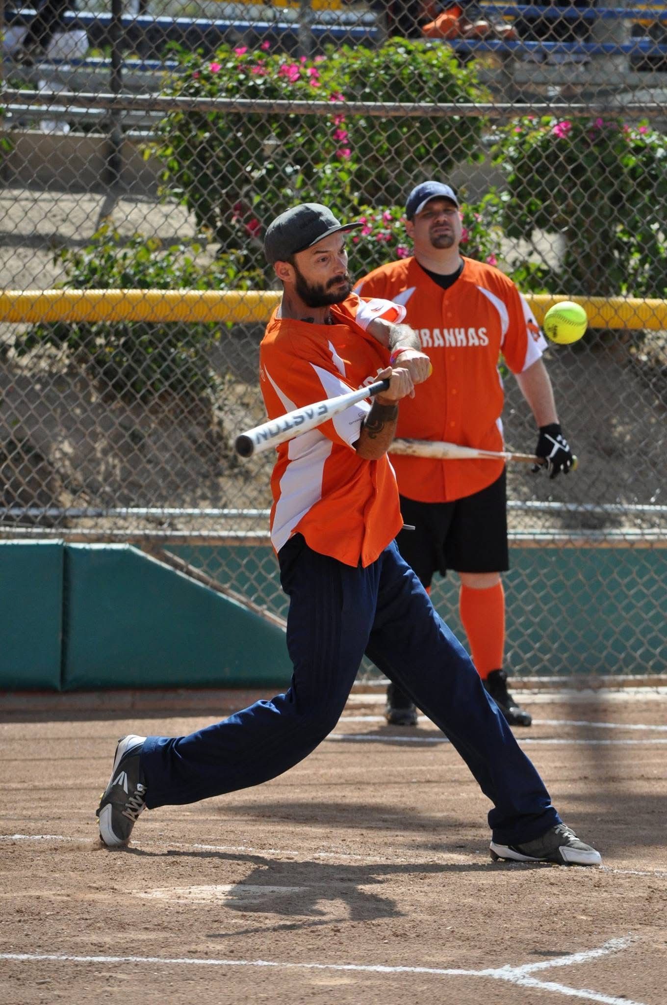 Baseball batter in orange swings at a pitch while a catcher stands behind home plate.