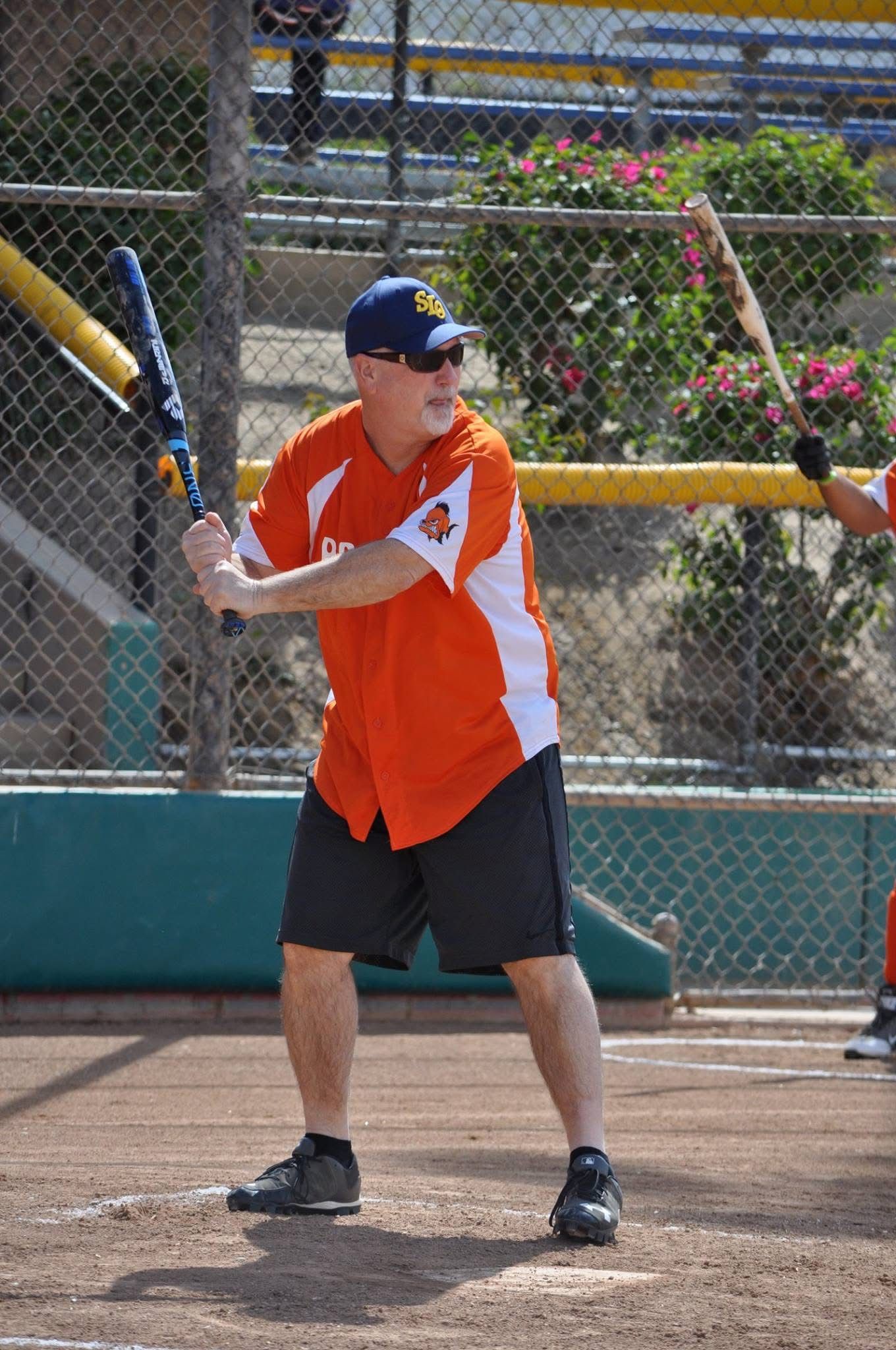 Softball player in orange jersey and blue cap preparing to swing on a dirt field