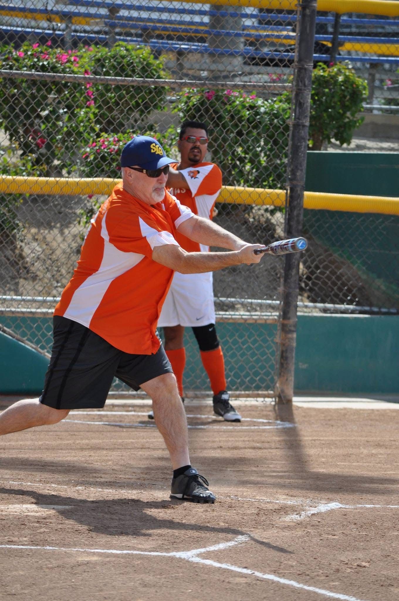 Two softball players in orange jerseys at bat on a dirt field, with a fence and bleachers behind them