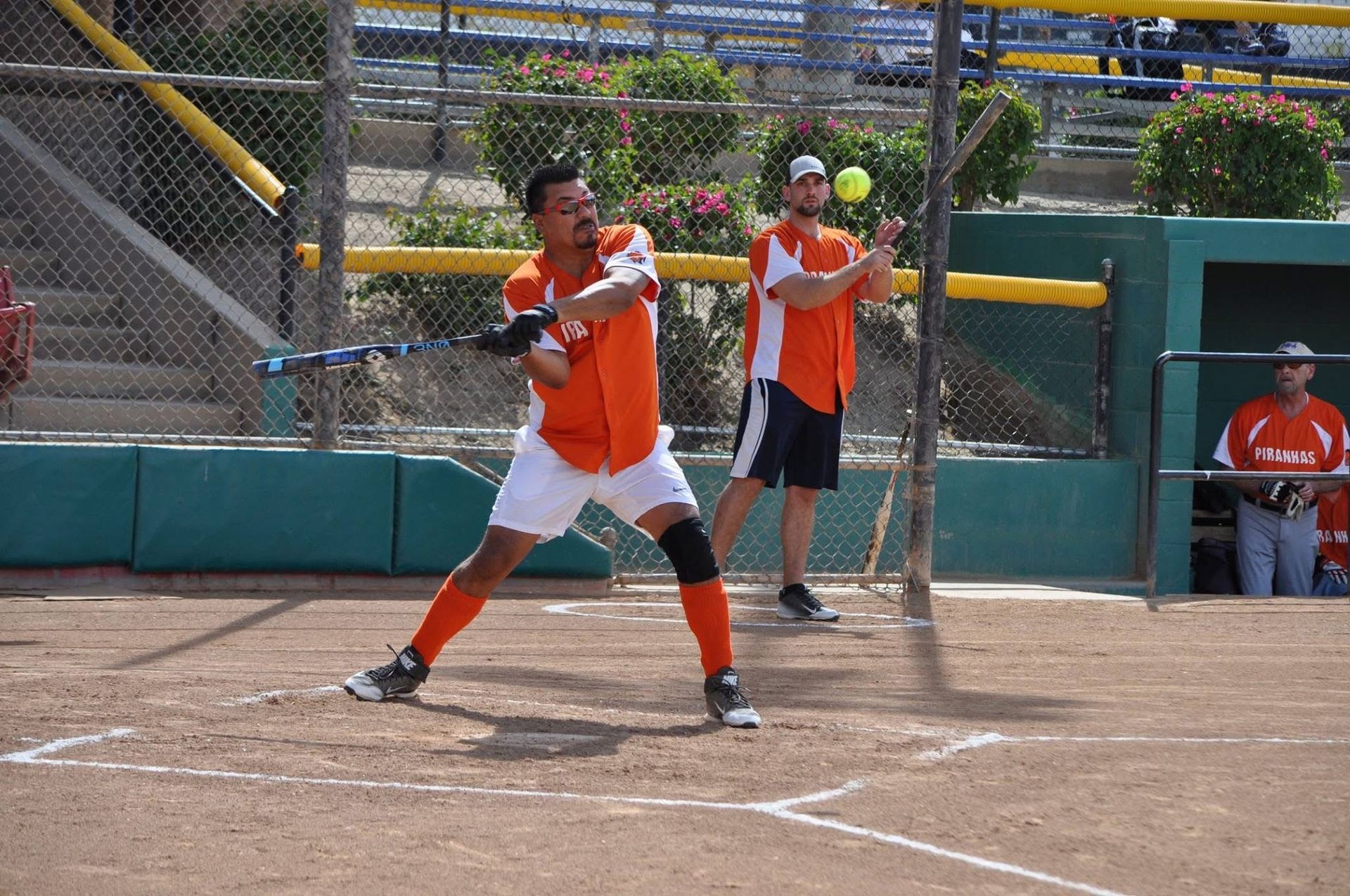 Softball batter in orange swings at home plate while catcher and umpire stand behind in a dirt field