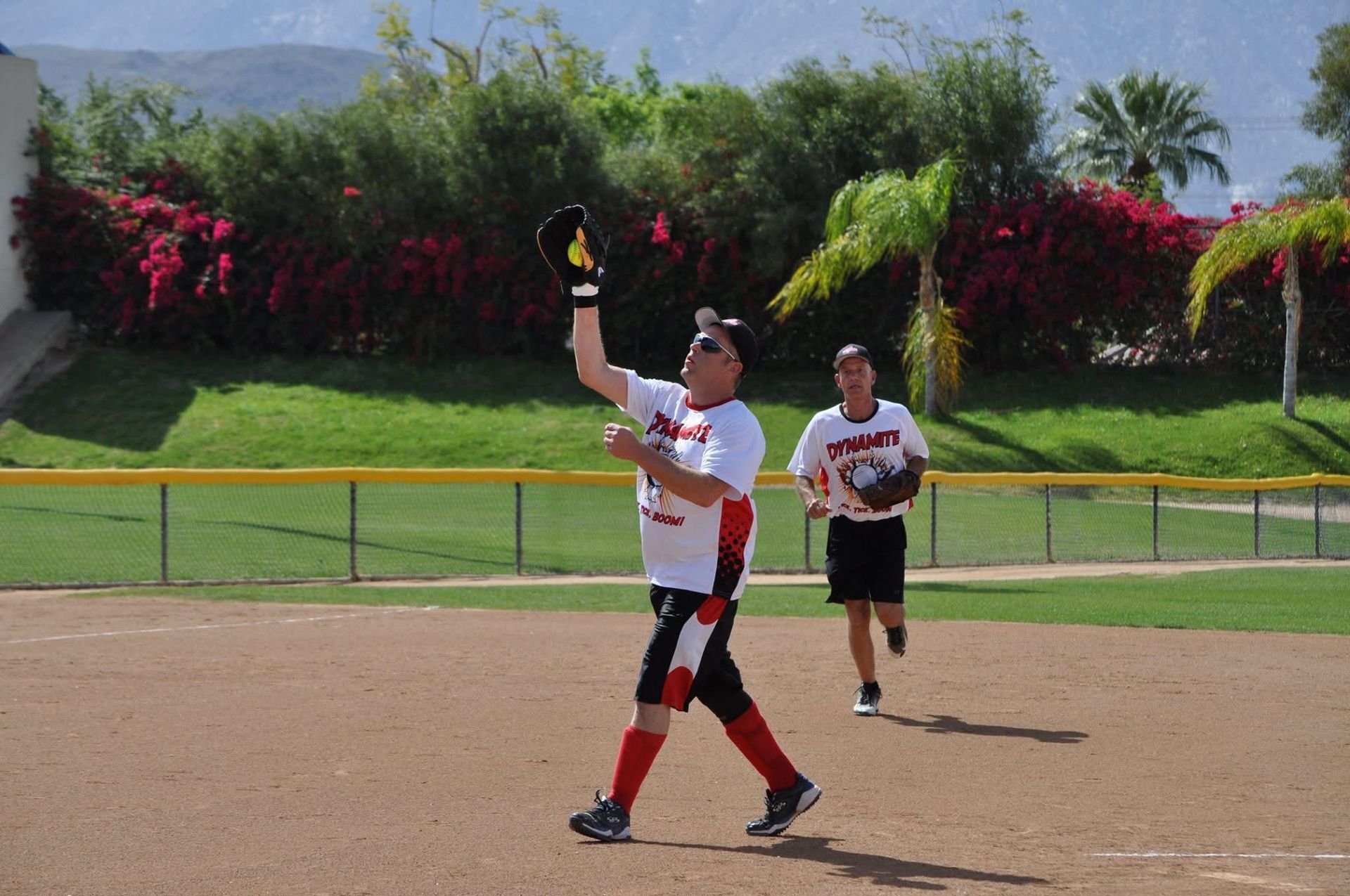 Softball player catches a ball under a sunny sky while a teammate runs behind her on the field.