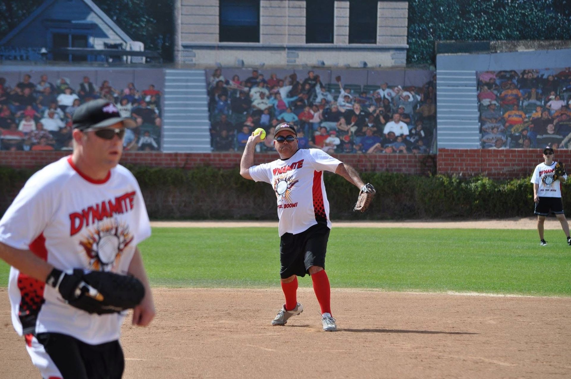 Baseball players on a sunny field, one pitching and another in the foreground near the mound.