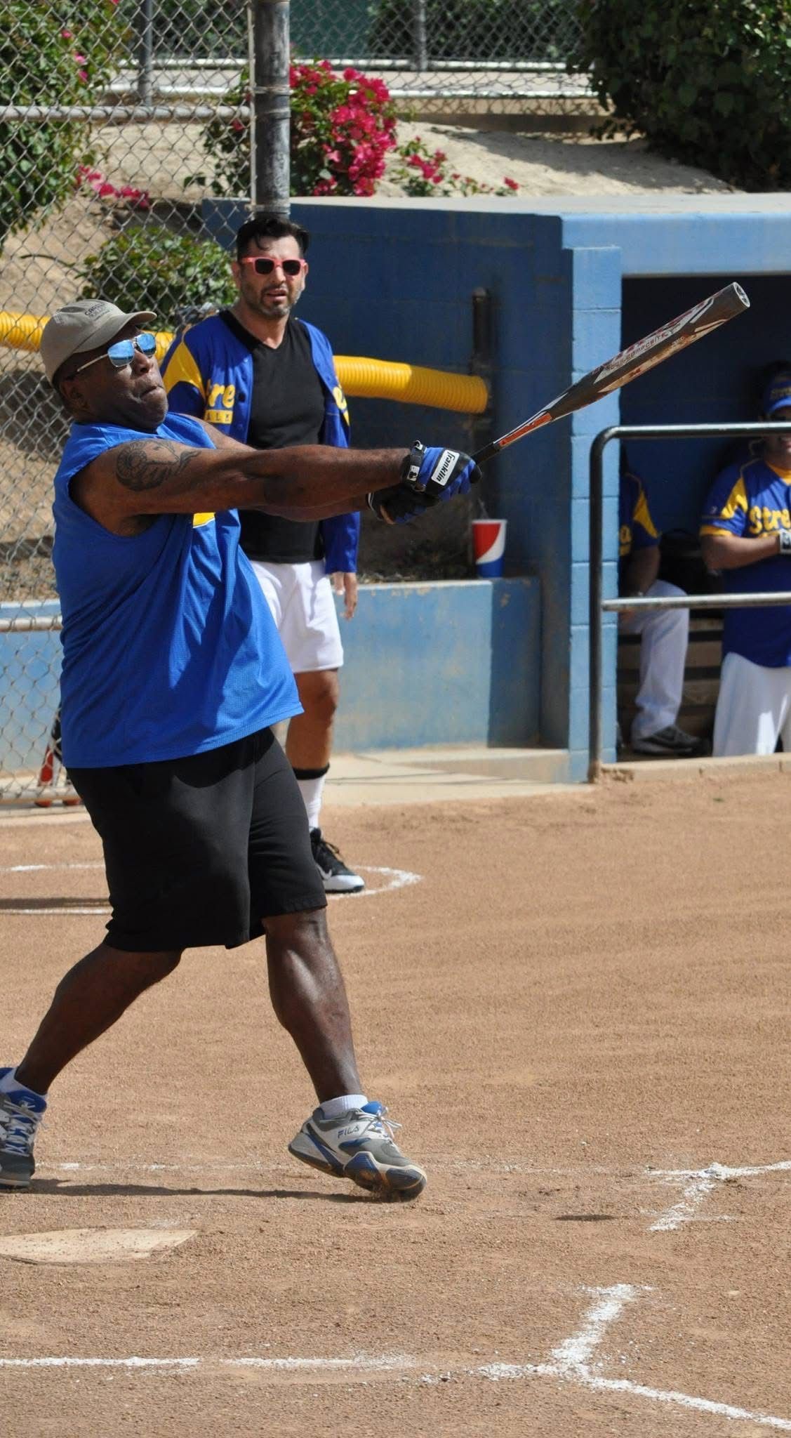 Softball batter in blue jersey swings at a pitch on a dirt field, with teammates visible in the dugout.