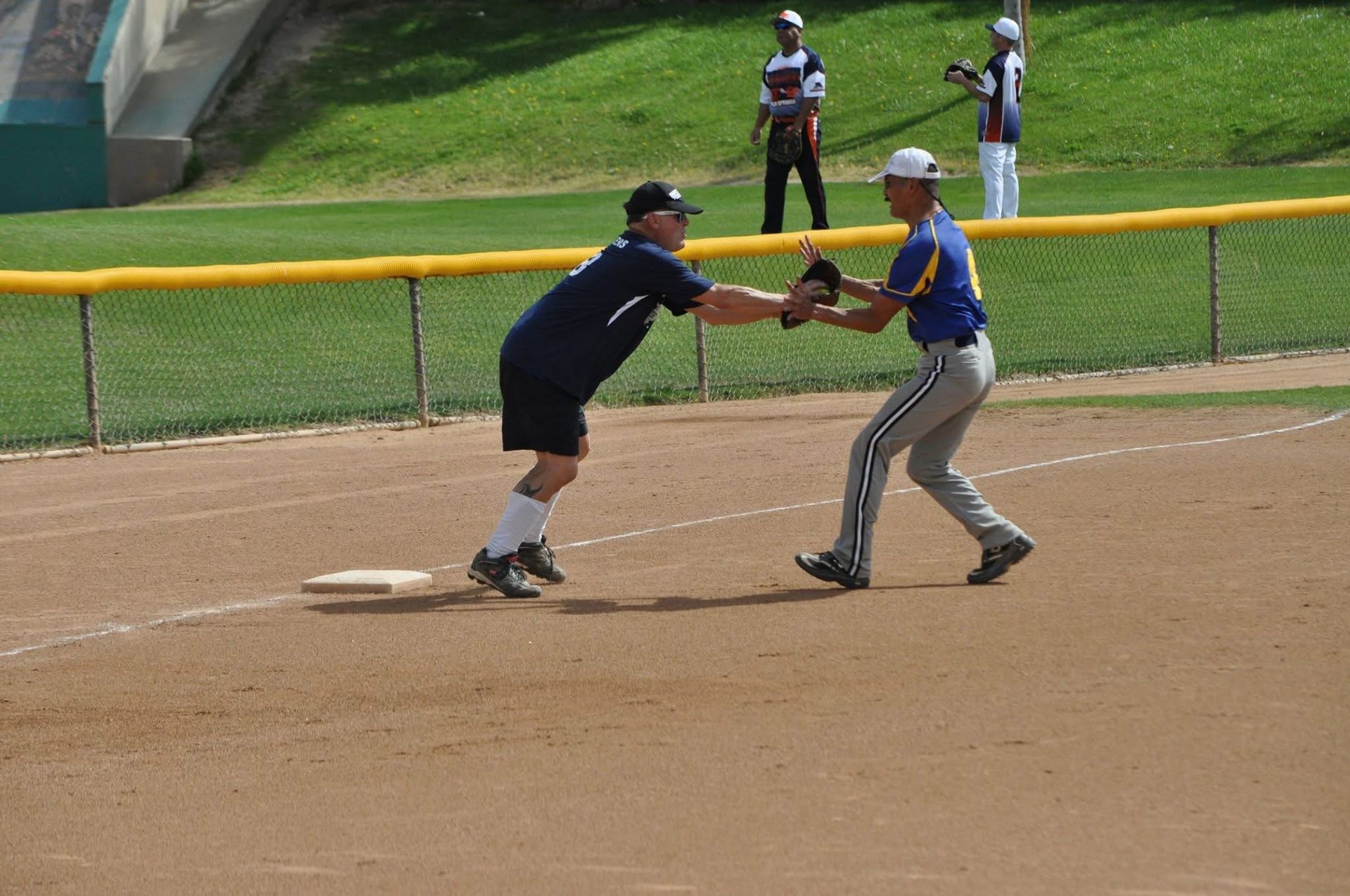 Two baseball players reach toward each other near first base on a dirt field, with outfielders in the background