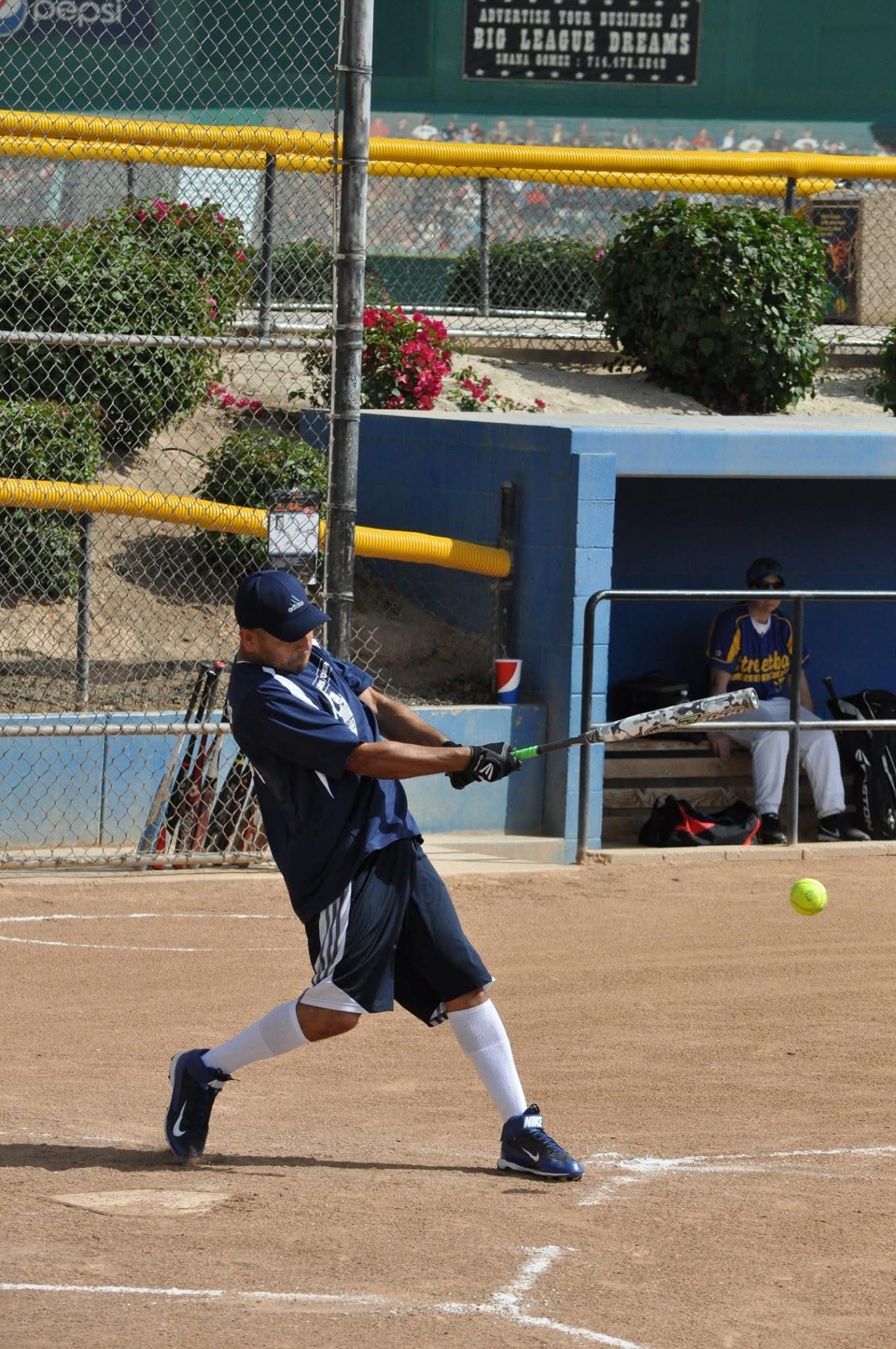 Softball player in blue uniform swings a bat on a dirt field, with a dugout and chain-link fence behind.