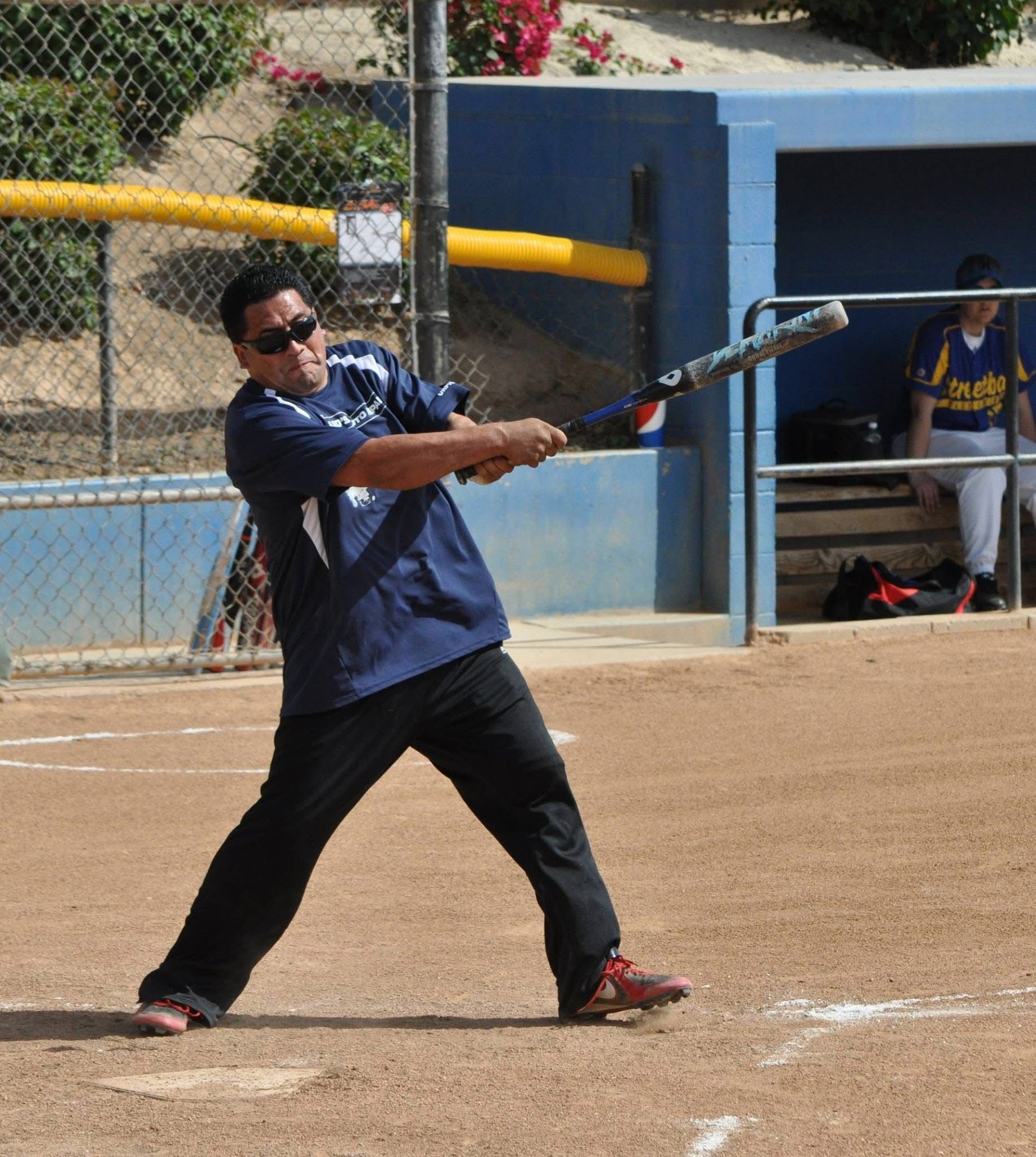 Baseball player in blue swings a bat on a dirt field near a dugout
