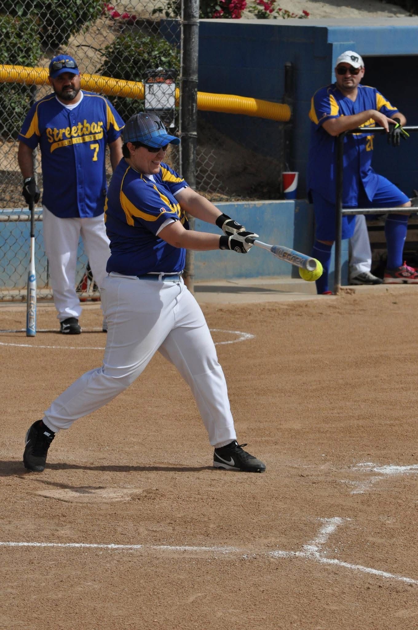 Softball batter in blue and yellow swings at a pitch on a dirt field, teammates watching nearby.