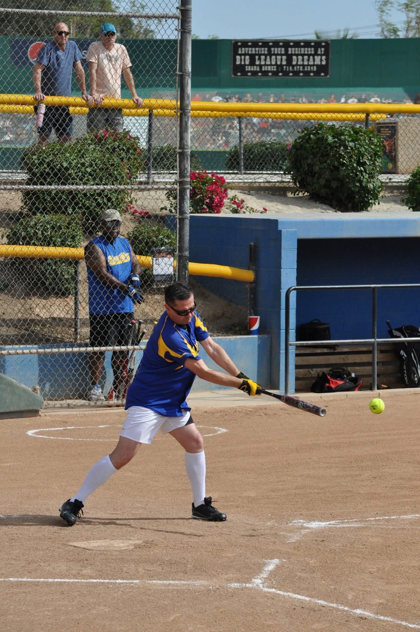 Softball player in blue and white swings at a pitch on a dirt field near the dugout.