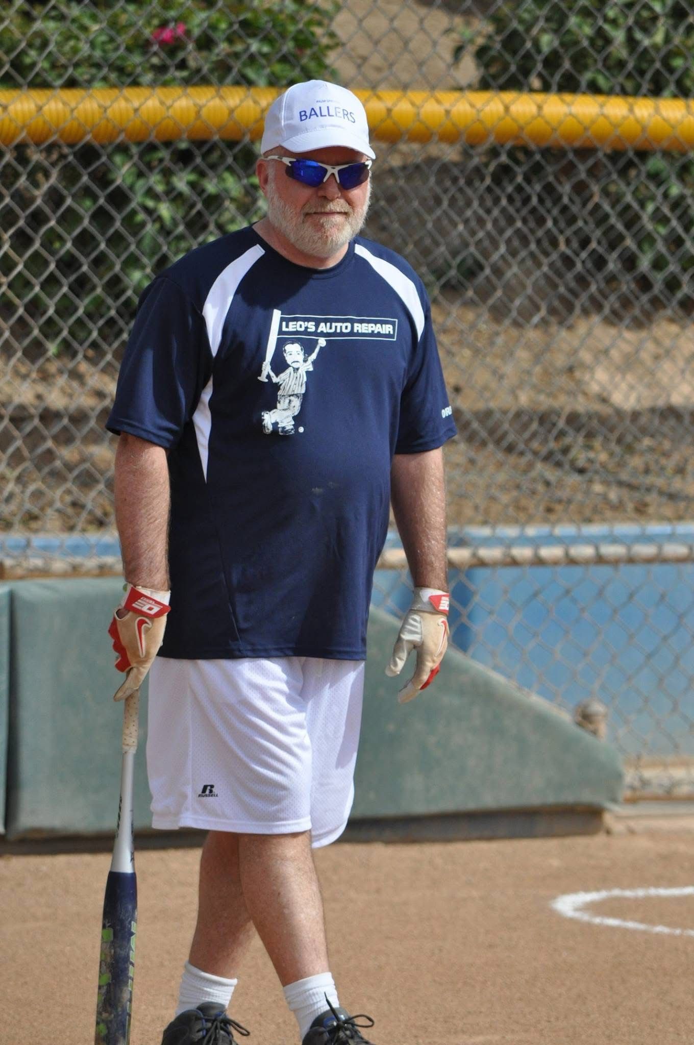 Man standing on a sports field in a navy jersey, white shorts, and cap, near a chain-link fence.